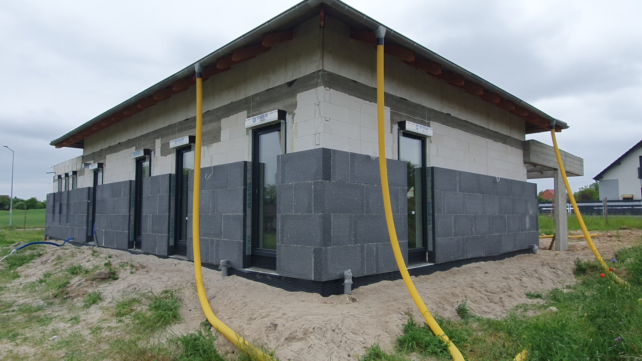 Niedokończona elewacja domu jednorodzinnego z częściowo nałożoną ciemnoszarą izolacją przyziemia, widoczne okna z roletami i żółte rury odprowadzające wodę deszczową.