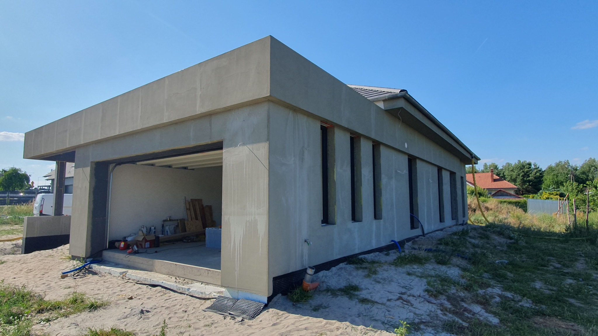 Surowy budynek z garażem w trakcie tynkowania elewacji, widoczne czarne obramowania okien, teren wokół budynku pokryty piaskiem, niebieskie rury na zewnątrz.
