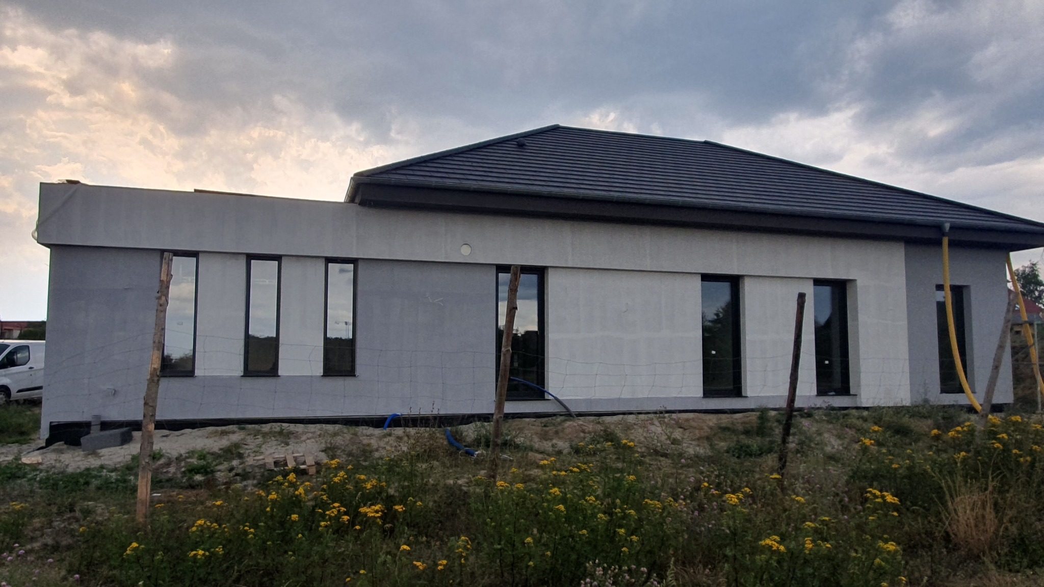 Dom jednorodzinny w trakcie prac wykończeniowych elewacji, widoczne fragmenty z nałożonym tynkiem w odcieniach szarości i białe płyty izolacyjne, okna z czarnymi ramami, w tle pochmurne niebo...