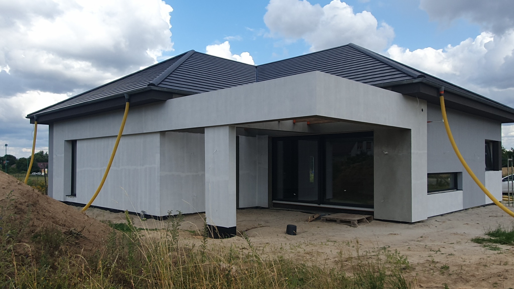 Surowa elewacja domu jednorodzinnego w trakcie prac wykończeniowych, widoczne żółte węże wentylacyjne i nierówny teren wokół budynku.