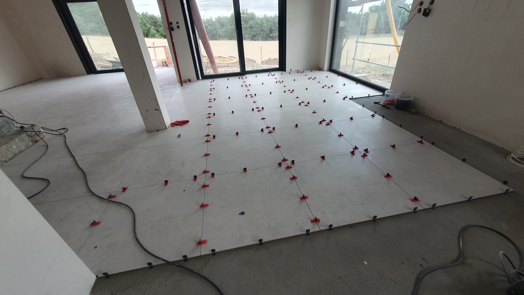 Układanie jasnych, kwadratowych płytek podłogowych w surowym wnętrzu, z użyciem czerwonych klinów i czarnych krzyżyków dystansowych, widoczne przewody i narzędzia.