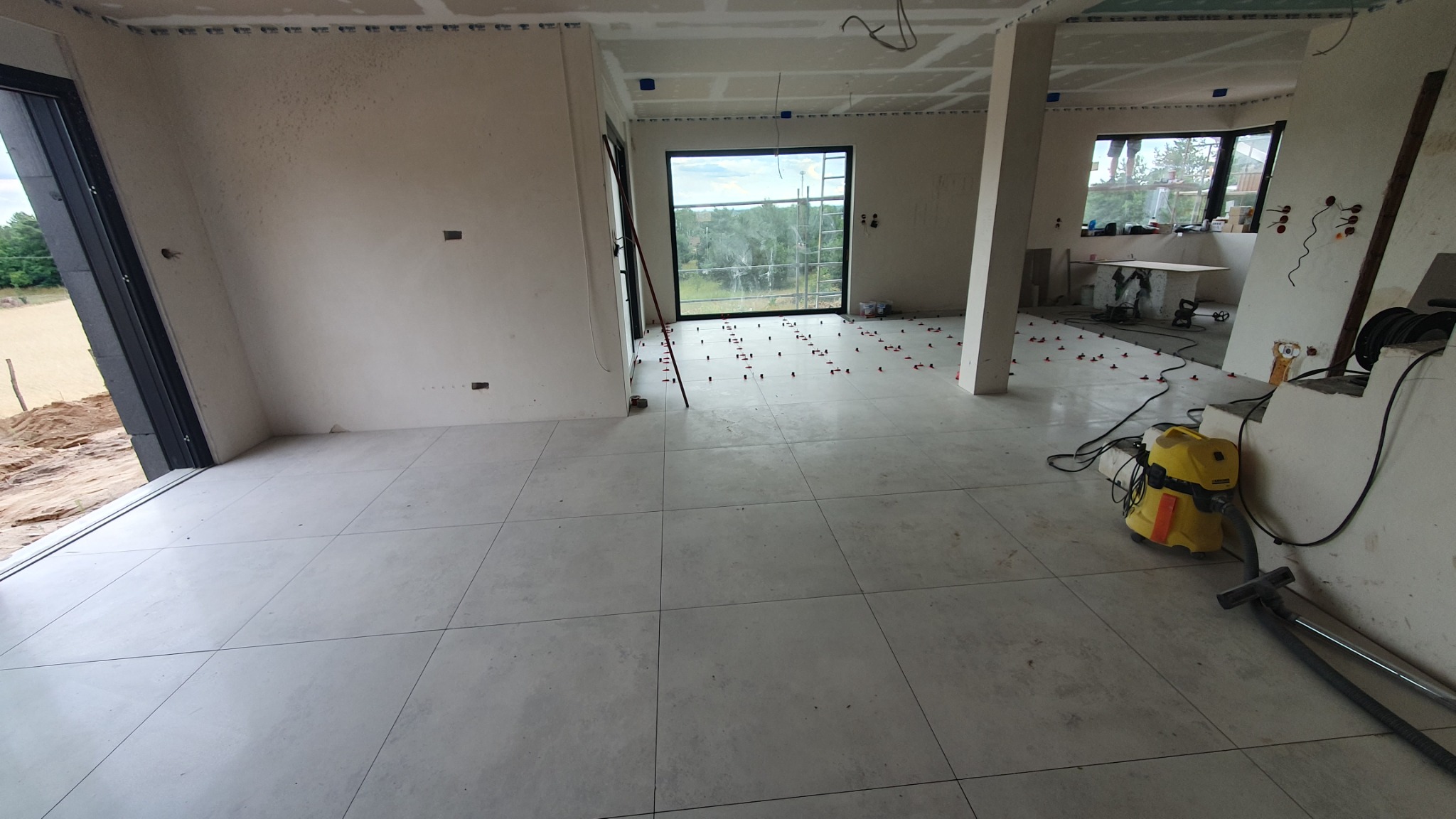Wnętrze w trakcie układania płytek podłogowych, z widocznymi krzyżykami dystansowymi, odkurzaczem przemysłowym i surowymi ścianami przygotowanymi do dalszych prac wykończeniowych.