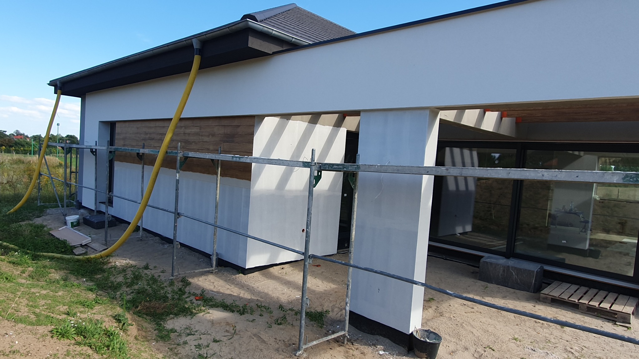 Biała elewacja domu jednorodzinnego w trakcie prac wykończeniowych, widoczne rusztowania, żółte węże do natrysku, fragment drewnianej okładziny i duże okna.