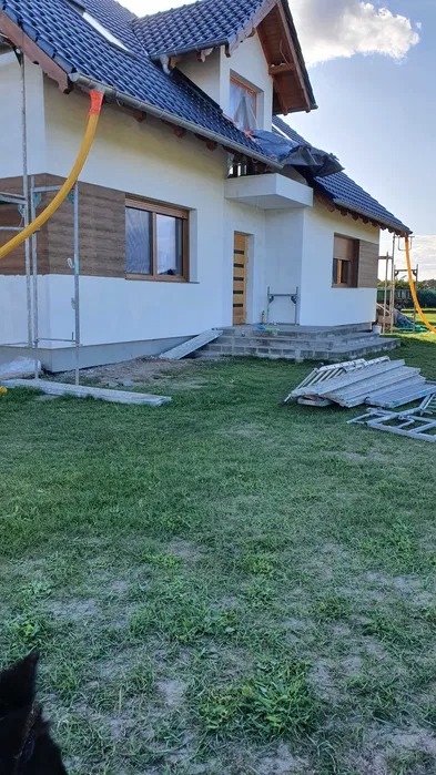 Dom jednorodzinny w trakcie prac elewacyjnych: częściowo ocieplona ściana z widoczną warstwą izolacyjną, rusztowanie z żółtym wężem, przygotowane schody wejściowe i materiały budowlane na trawniku.