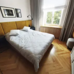 Sypialnia z żółtym panelem za łóżkiem, drewnianą podłogą w jodełkę i oknem z drewnianymi lamelami.