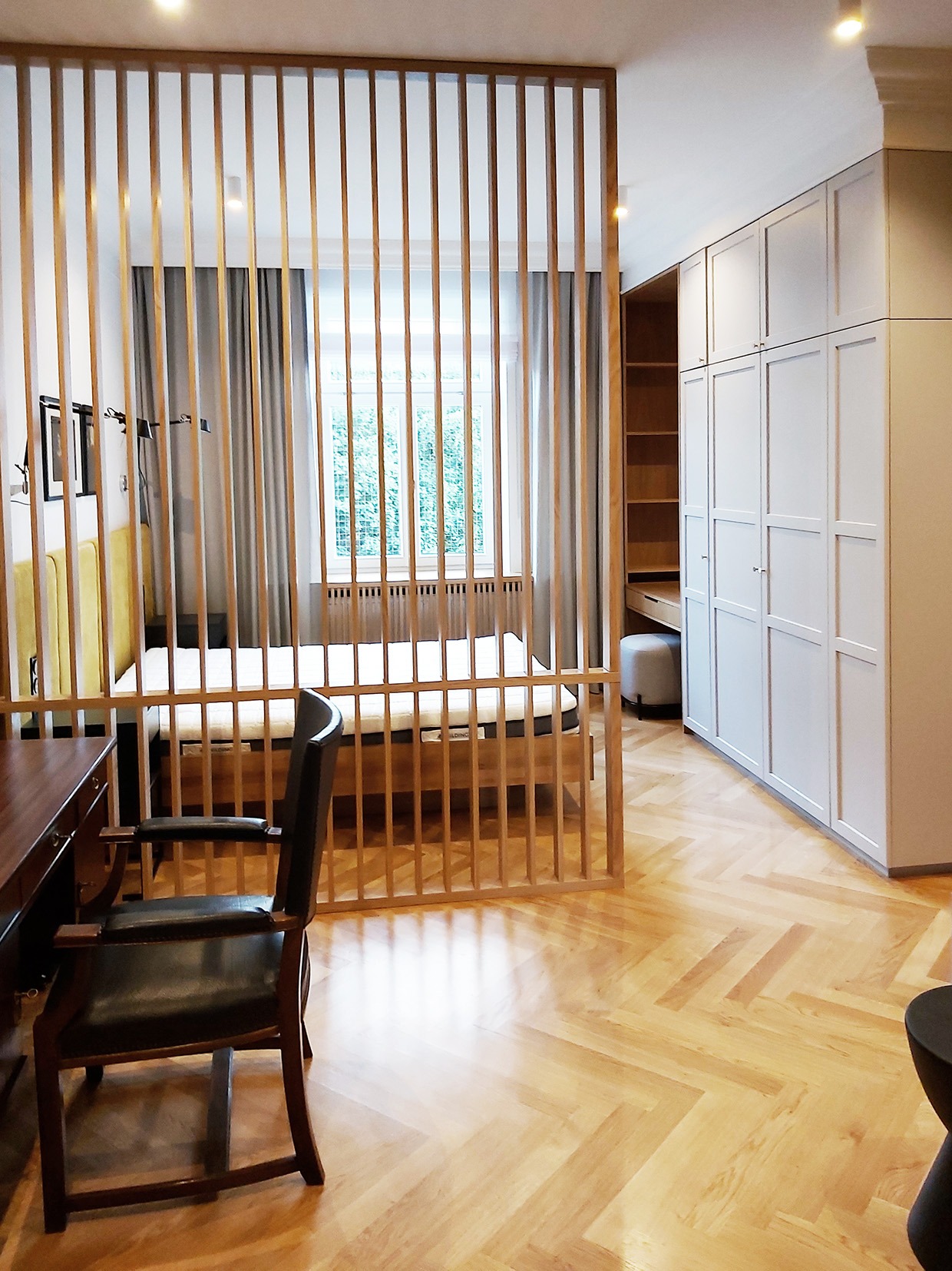 Pokój z drewnianą ażurową ścianką działową, łóżkiem z żółtym zagłówkiem, biurkiem z krzesłem i szafą wnękową z białymi frontami oraz parkietem ułożonym w jodełkę.