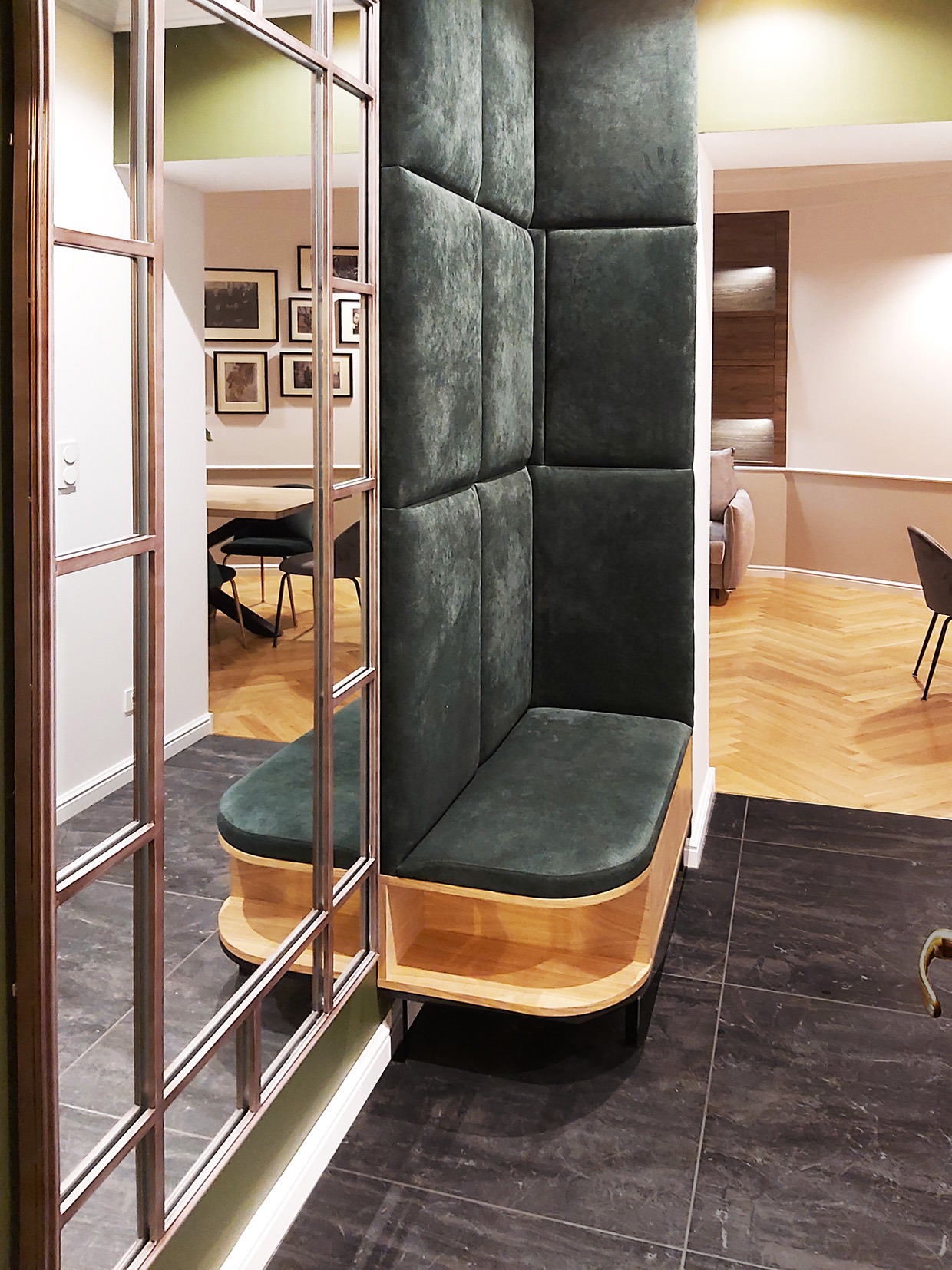 Wysoka, tapicerowana na ciemnozielono narożna ławka wbudowana w ścianę, z drewnianą podstawą, widziana przez szklaną ściankę działową w nowoczesnym wnętrzu z ciemną podłogą i jasnym parkietem...