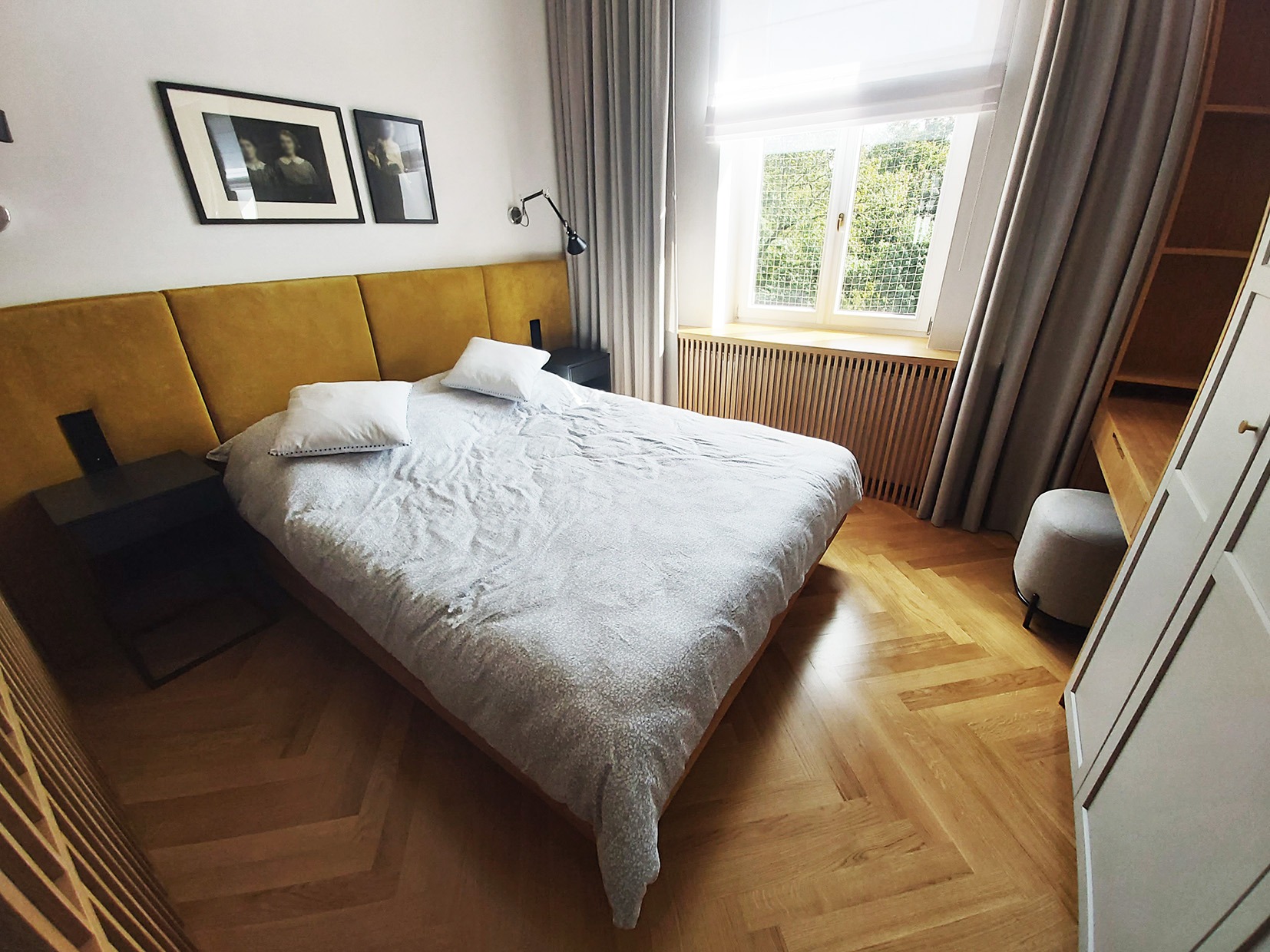 Sypialnia z żółtym panelem za łóżkiem, drewnianą podłogą w jodełkę i oknem z drewnianymi lamelami.