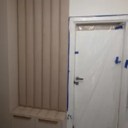Beżowy panel ścienny z tapicerowanym siedziskiem i szafkami, obok drzwi zabezpieczonych folią malarską.