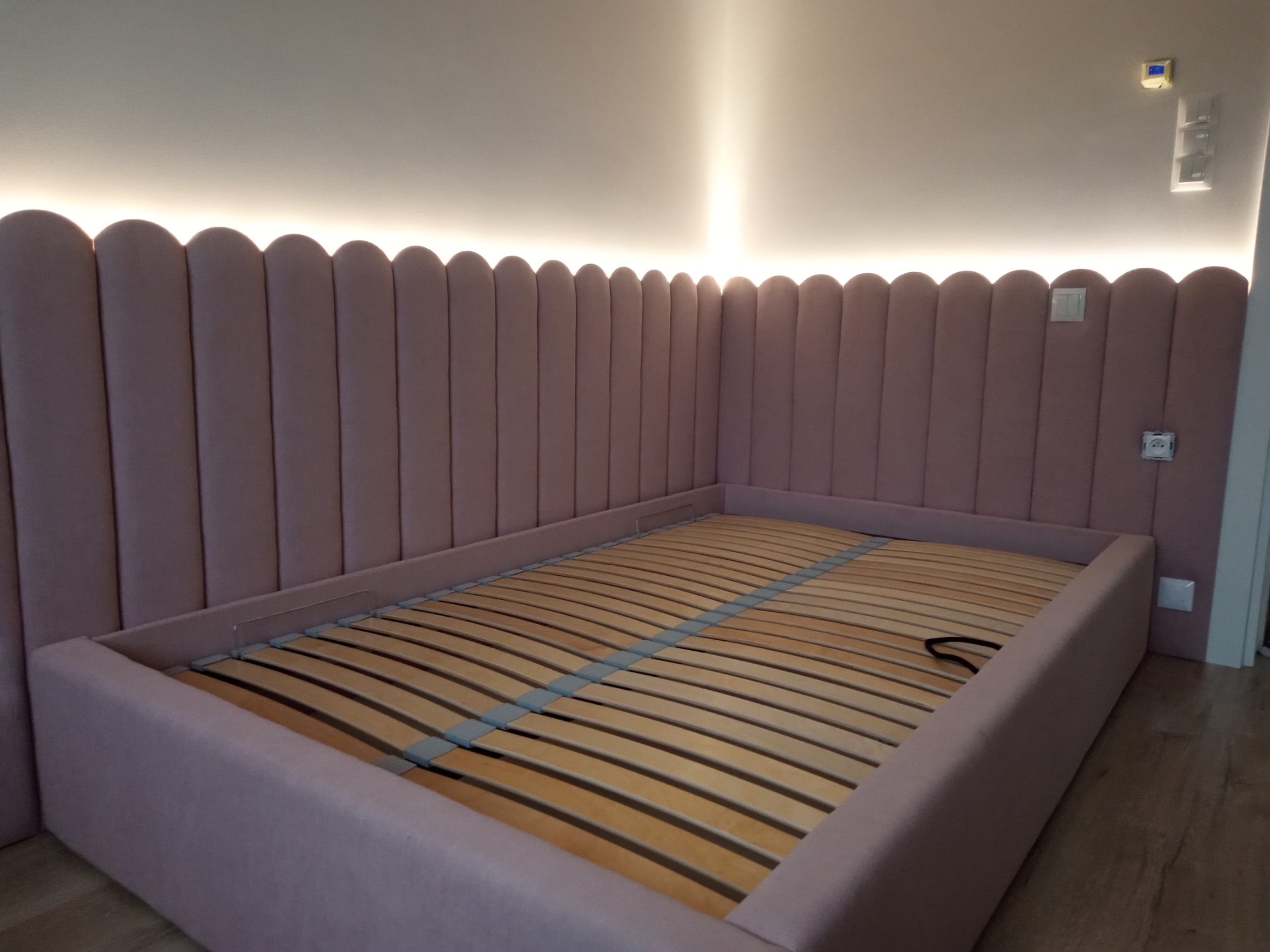 Różowe tapicerowane łóżko z wezgłowiem w kształcie zaokrąglonych paneli, oświetlone od góry, z drewnianym stelażem bez materaca, widoczne gniazdka elektryczne na ścianie.