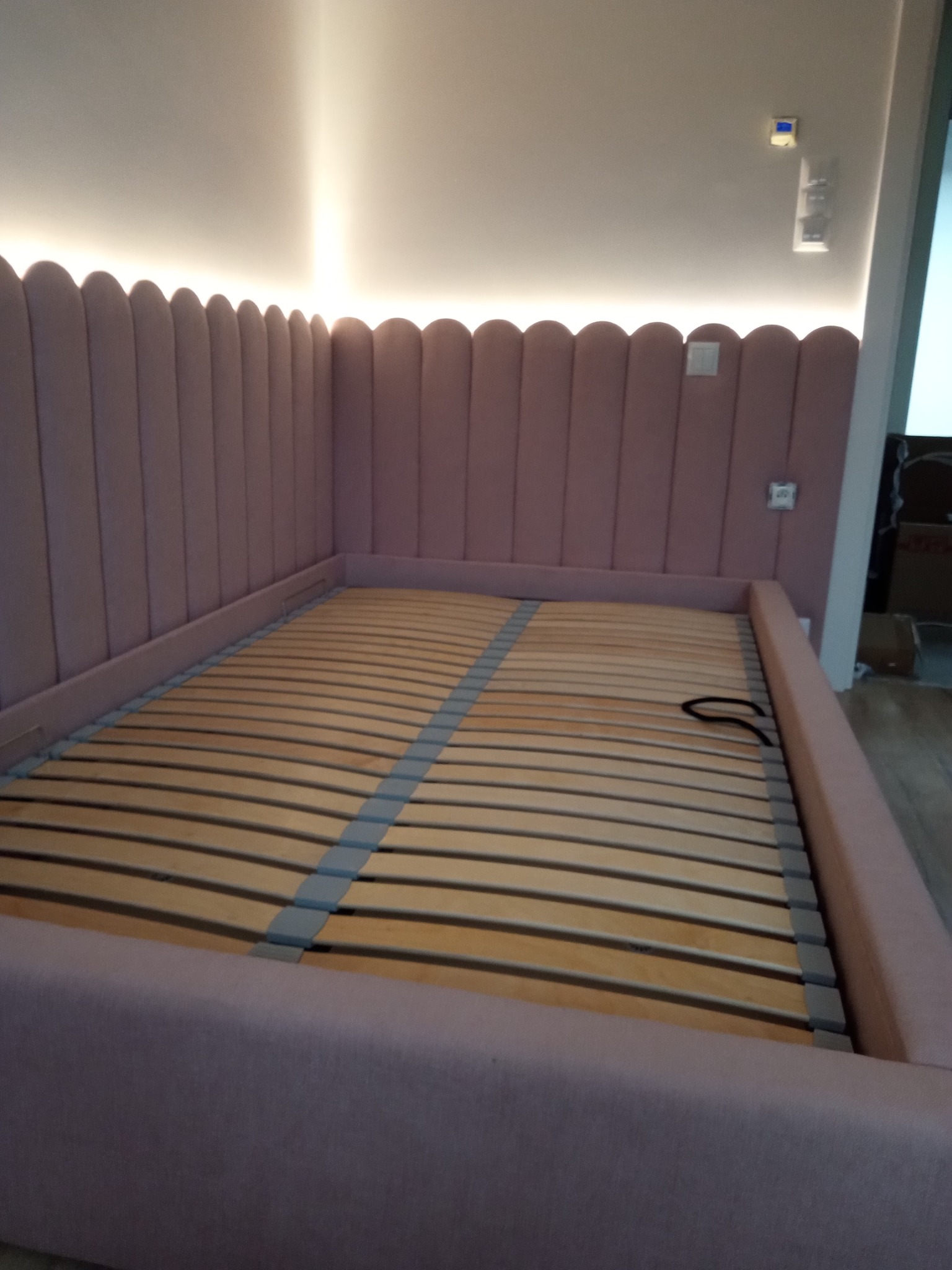 Różowe łóżko z tapicerowanym wezgłowiem w kształcie muszelek i drewnianym stelażem, oświetlone od góry, w jasnym wnętrzu.