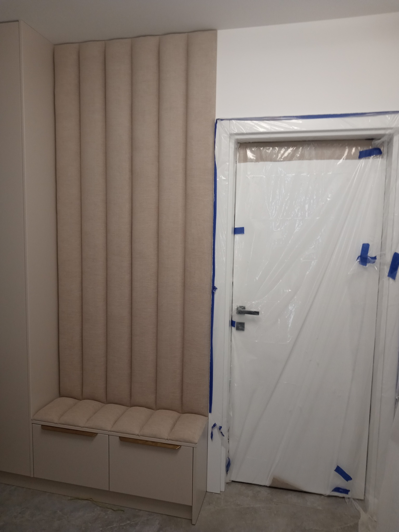 Beżowy panel ścienny z tapicerowanym siedziskiem i szafkami, obok drzwi zabezpieczonych folią malarską.