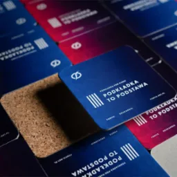 Kwadratowe podkładki pod kubek z logo firmy i hasłem reklamowym w kolorach niebieskim, czerwonym i korkowym, ułożone pod kątem na ciemnym tle.