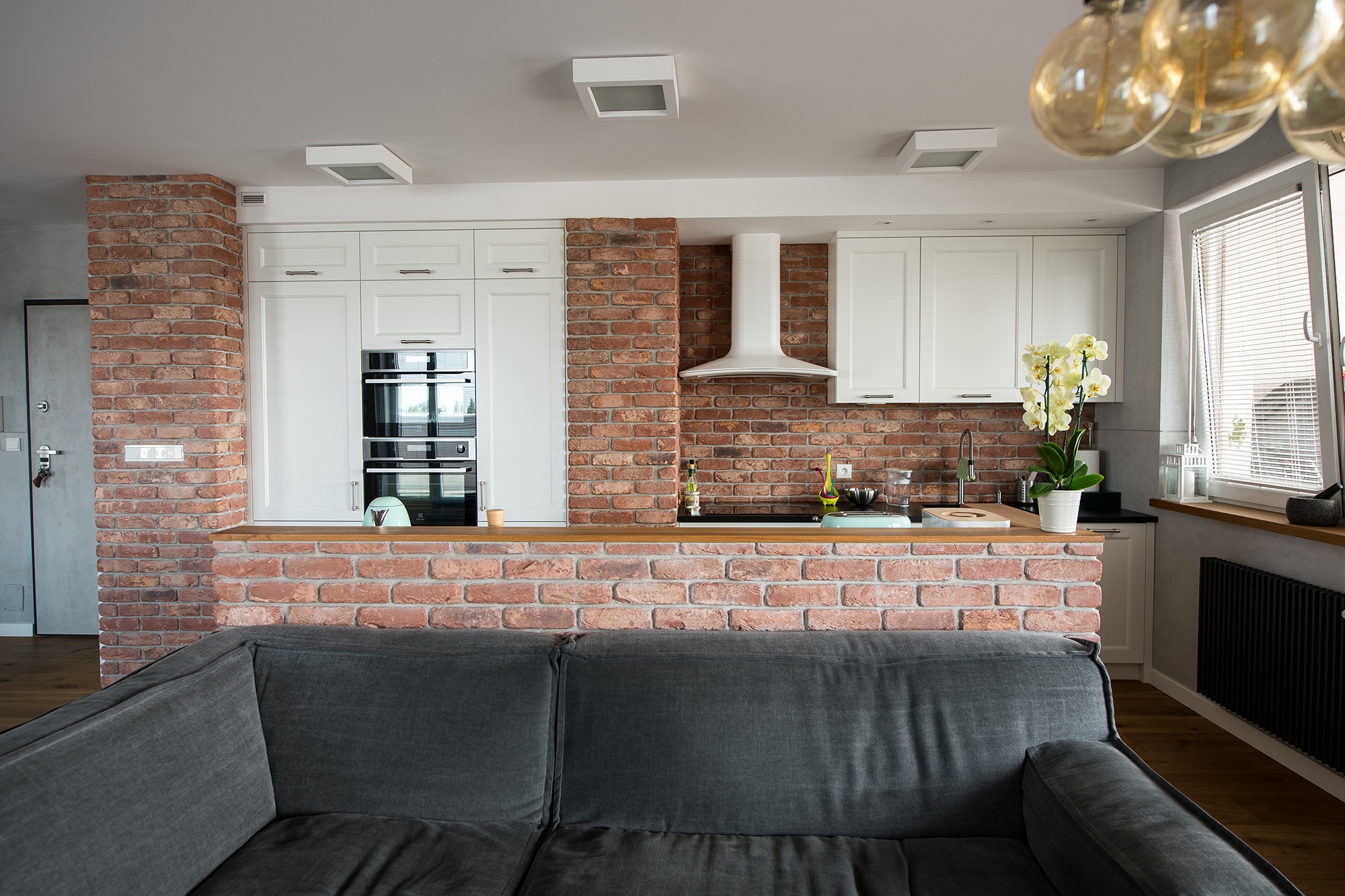 Salon z aneksem kuchennym w stylu loftowym: ceglana ściana, białe szafki kuchenne, szara sofa na pierwszym planie.