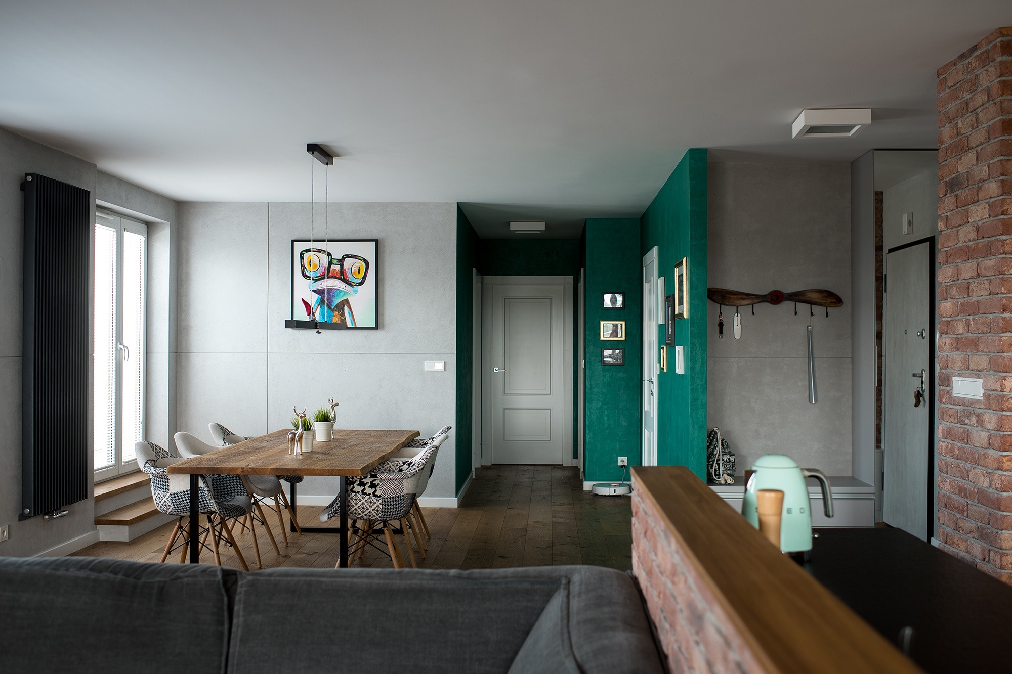 Wnętrze mieszkania z drewnianym stołem i krzesłami o wzorzystych obiciach, widok na przedpokój z zieloną ścianą i dekoracjami, w tle ściana z cegły.