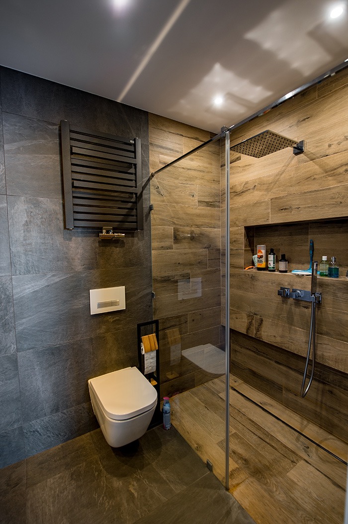 Nowoczesna łazienka z szarymi i drewnopodobnymi płytkami, kabiną prysznicową typu walk-in z deszczownicą, wiszącą toaletą i grzejnikiem drabinkowym.
