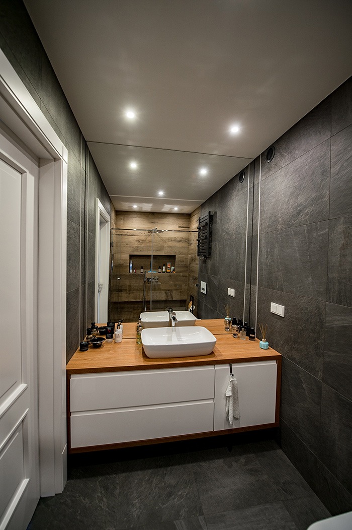 Nowoczesna łazienka z szarymi płytkami, drewnianym blatem pod umywalką nablatową i dużym lustrem, w którym odbija się kabina prysznicowa z drewnianymi akcentami.