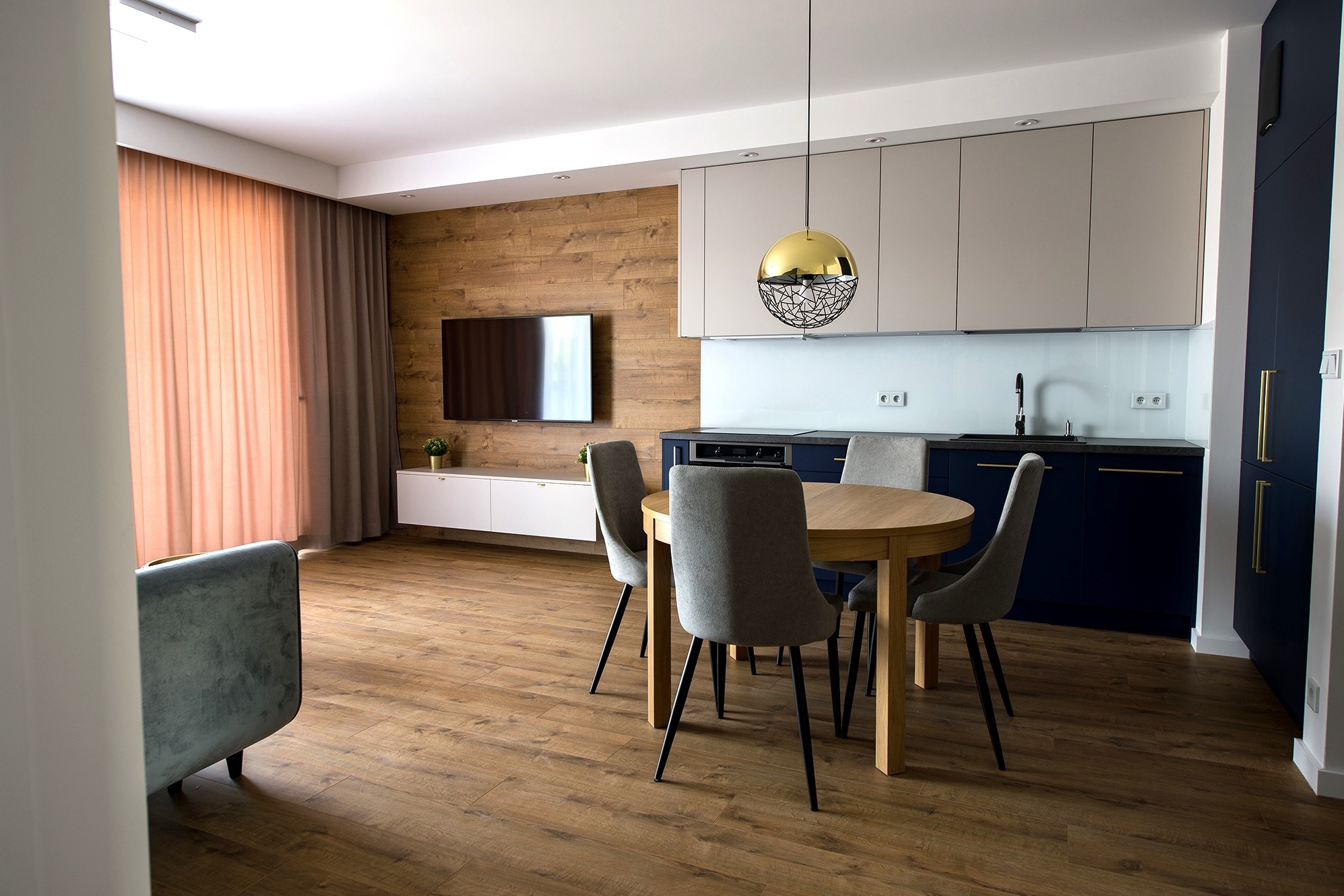 Nowoczesne wnętrze: okrągły drewniany stół z szarymi krzesłami, złota lampa wisząca nad stołem, drewniana ściana z telewizorem, biała szafka RTV, niebieska zabudowa kuchenna z beżowymi szafkami...