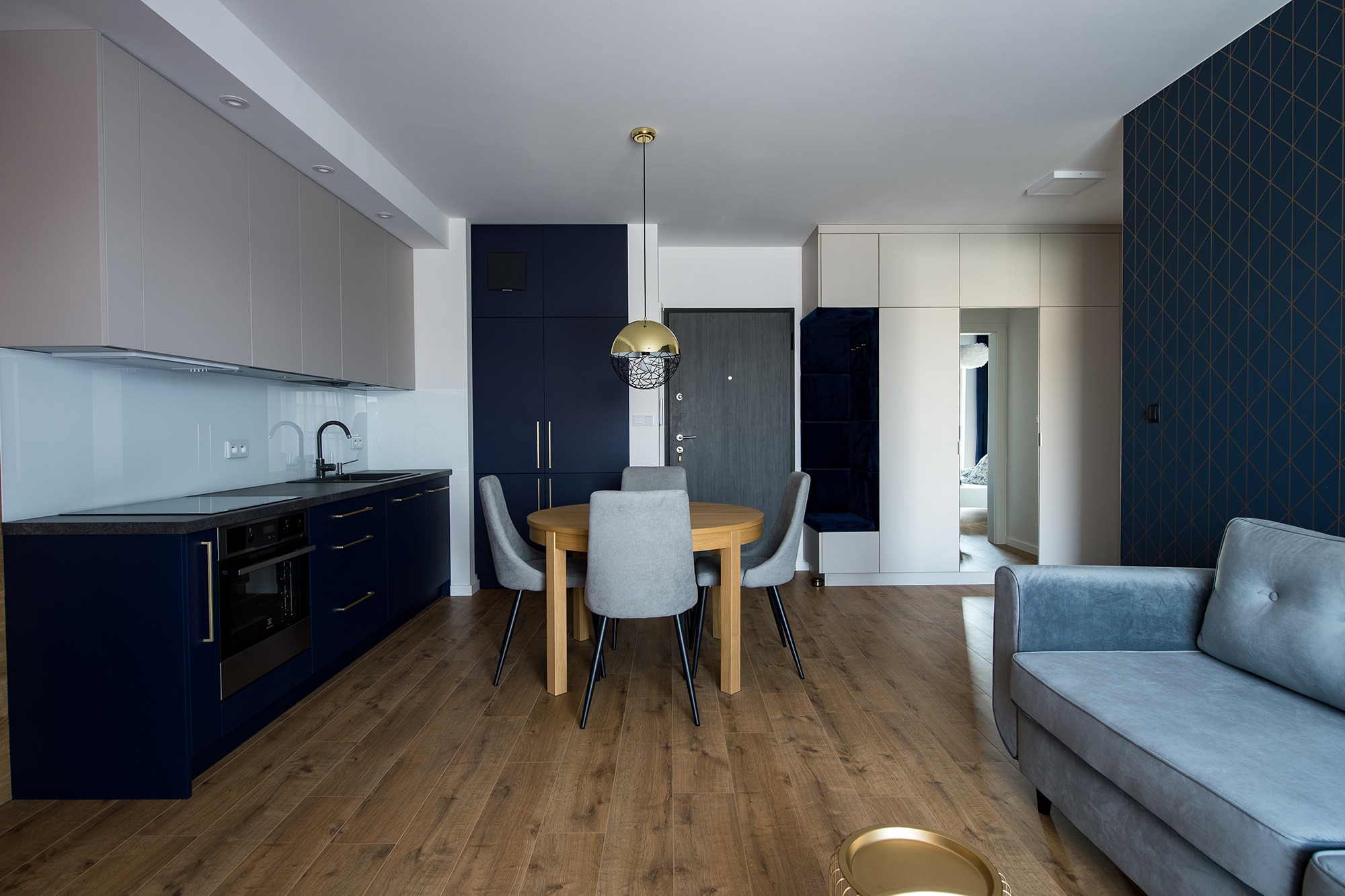 Nowoczesne wnętrze mieszkania: drewniana podłoga, okrągły stół z szarymi krzesłami, niebieska zabudowa kuchenna z piekarnikiem, szare szafki wiszące, niebieska ściana z geometrycznym wzorem, szara...