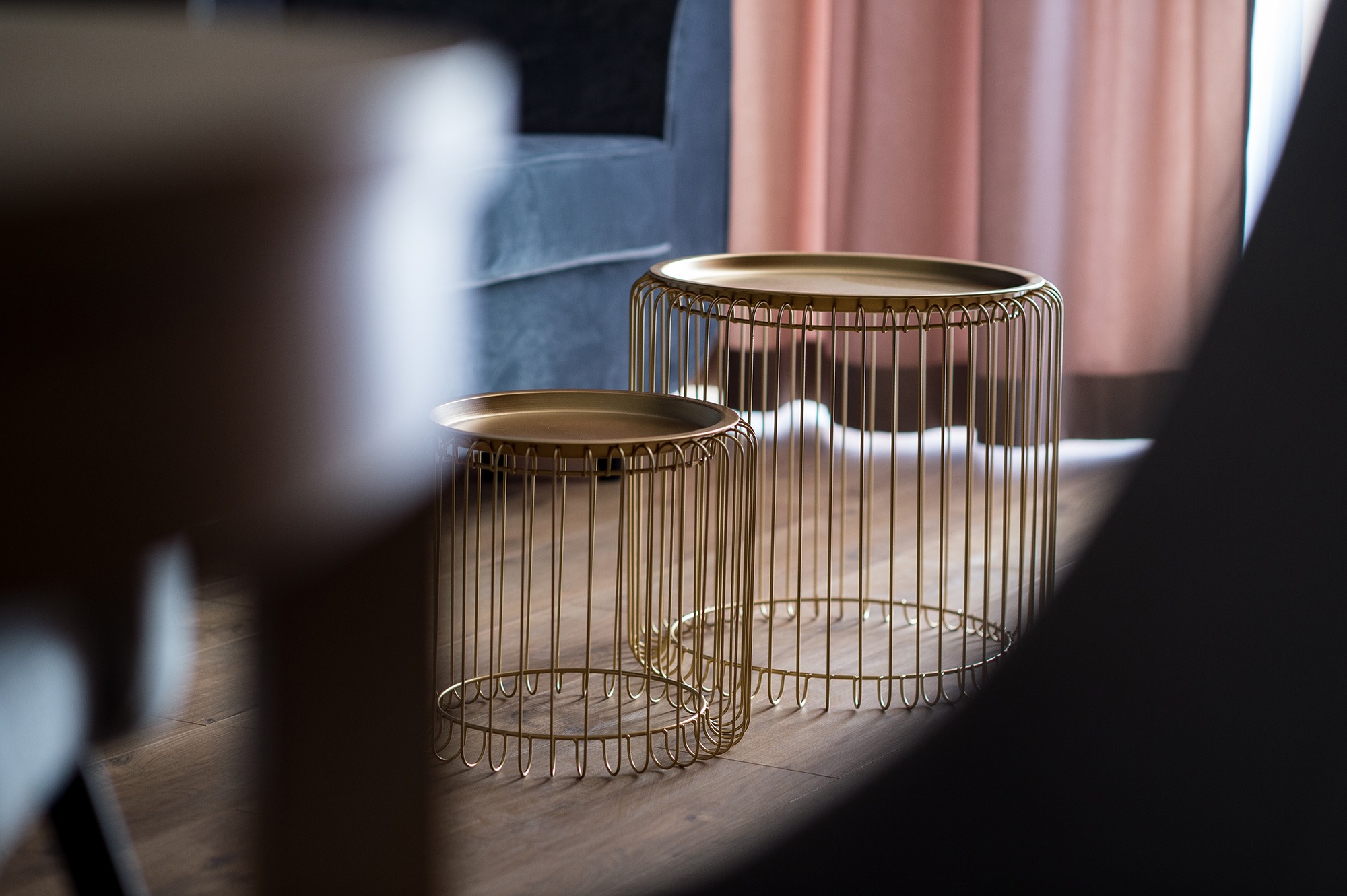 Dwa złote stoliki kawowe z metalowych prętów na drewnianej podłodze, z widocznymi elementami sofy i zasłon w tle.