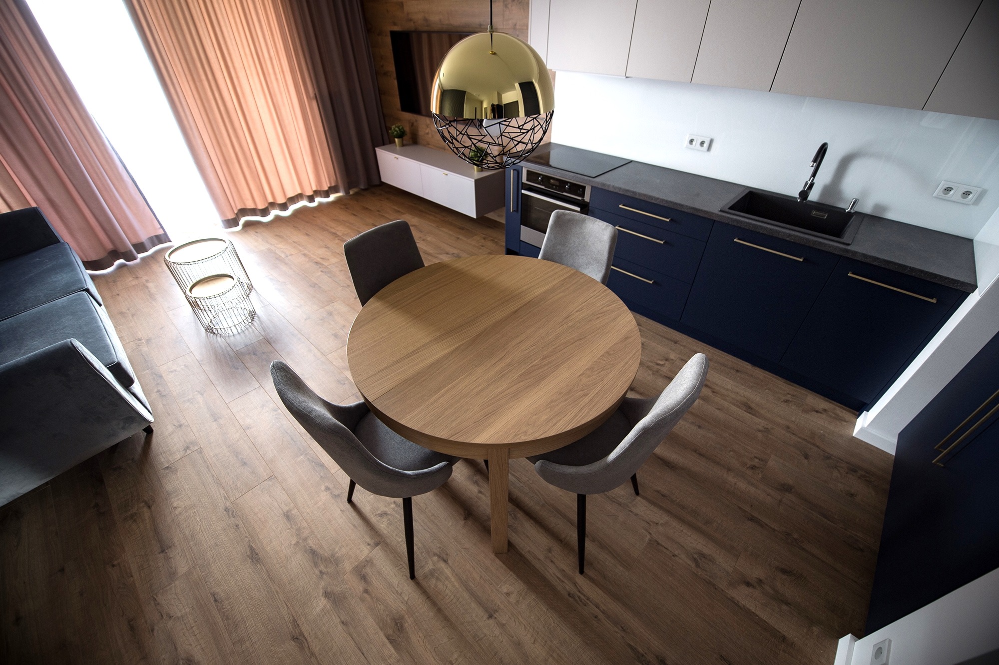 Nowoczesna aranżacja wnętrza z okrągłym drewnianym stołem, szarymi krzesłami i granatową zabudową kuchenną z złotymi uchwytami, widok z góry.