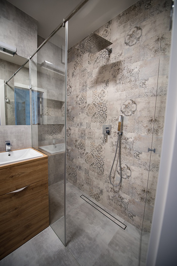 Nowoczesna łazienka z kabiną prysznicową typu walk-in, deszczownicą i ścianą wyłożoną patchworkowymi płytkami w odcieniach beżu i szarości, widoczny odpływ liniowy w podłodze.