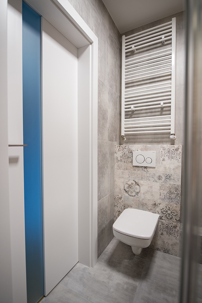 Nowoczesna łazienka z podwieszaną toaletą, grzejnikiem drabinkowym i drzwiami z niebieskim akcentem.