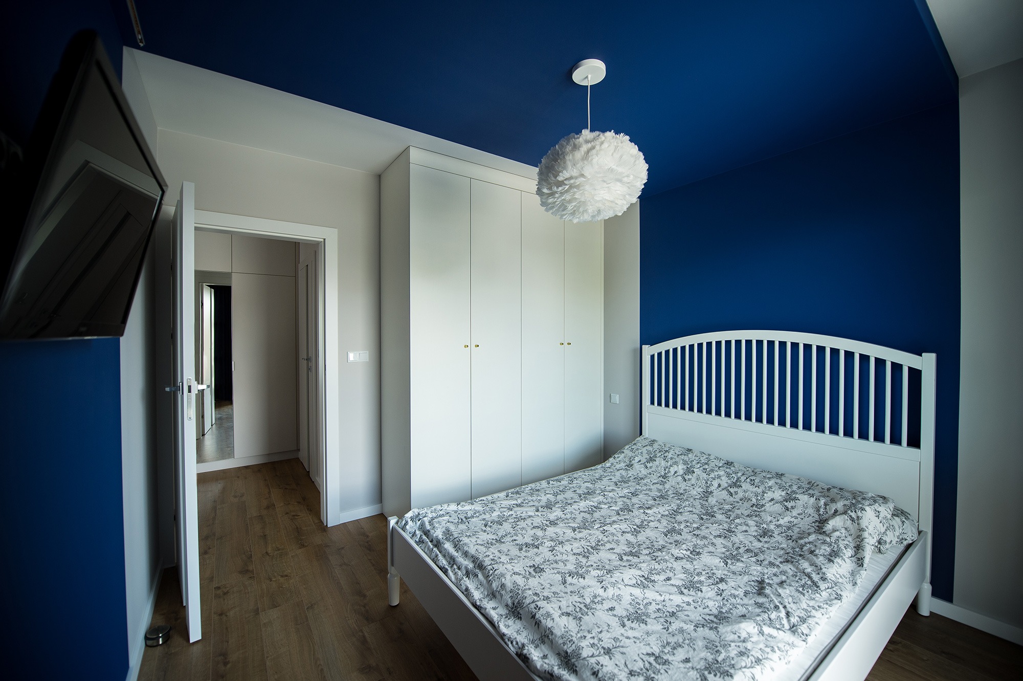 Nowoczesna sypialnia z białym łóżkiem, szafą w zabudowie i intensywnie niebieskim sufitem, oświetlona kulistą lampą z piór. Widoczny otwarty pokój przechodzący do kolejnego pomieszczenia.