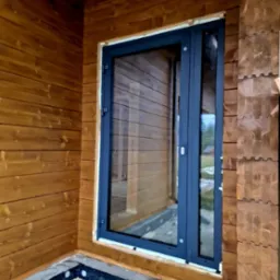 Nowe okno PCV w ciemnej ramie zamontowane w drewnianym domu z bali, widoczna piana montażowa i folia fundamentowa.