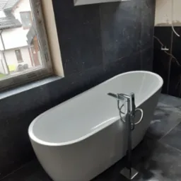 Nowoczesna, wolnostojąca wanna z chromowaną baterią w łazience z czarnymi, marmurkowymi płytkami, widok na okno dachowe i okno z widokiem na dom sąsiada.