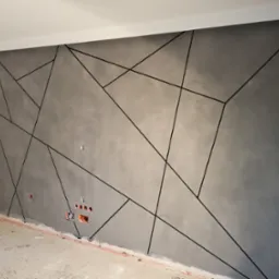 Ściana pokryta szarą gładzią z geometrycznym wzorem z czarnych linii, widoczne gniazdka elektryczne, surowa podłoga w trakcie prac wykończeniowych.