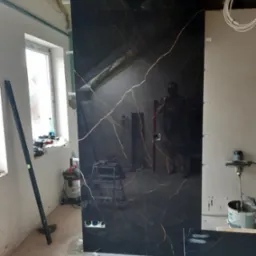 Montaż dużej płyty z czarnego gresu imitującego marmur z złotymi żyłami, widoczne przygotowane otwory na gniazdka elektryczne, poziomica oparta o ścianę, pomieszczenie w trakcie prac wykończeniowych.