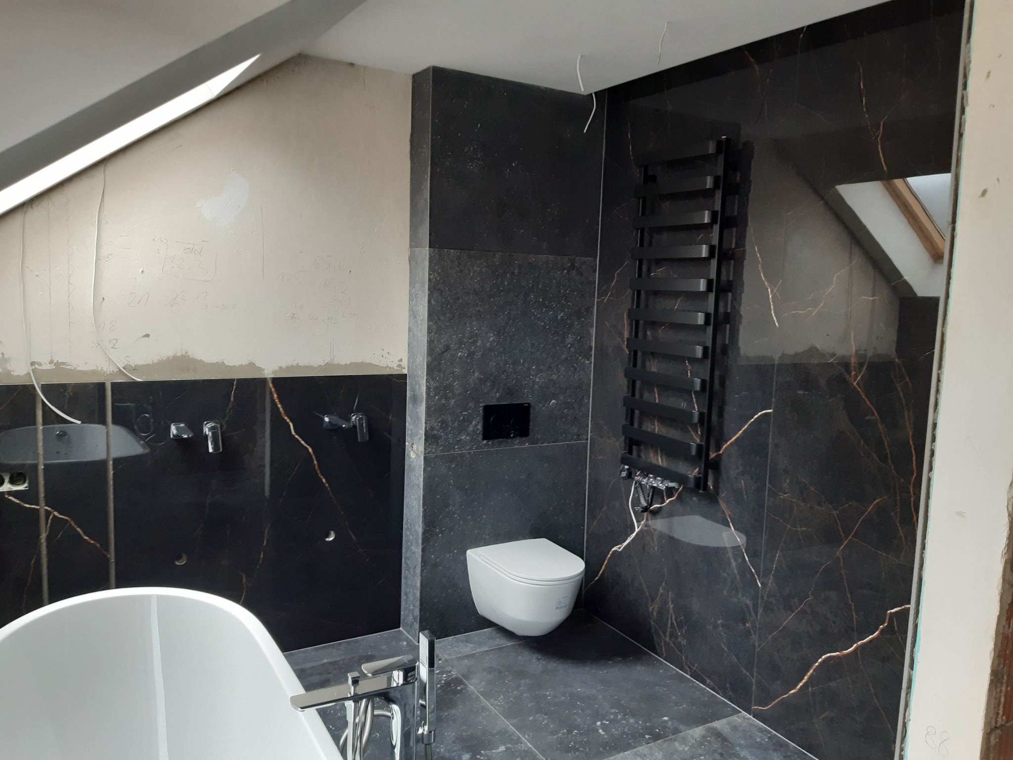 Łazienka w trakcie remontu: czarne płytki imitujące marmur z złotymi żyłami na ścianach, biała wisząca toaleta, grzejnik drabinkowy w kolorze czarnym, fragment wanny i ściana w stanie surowym...