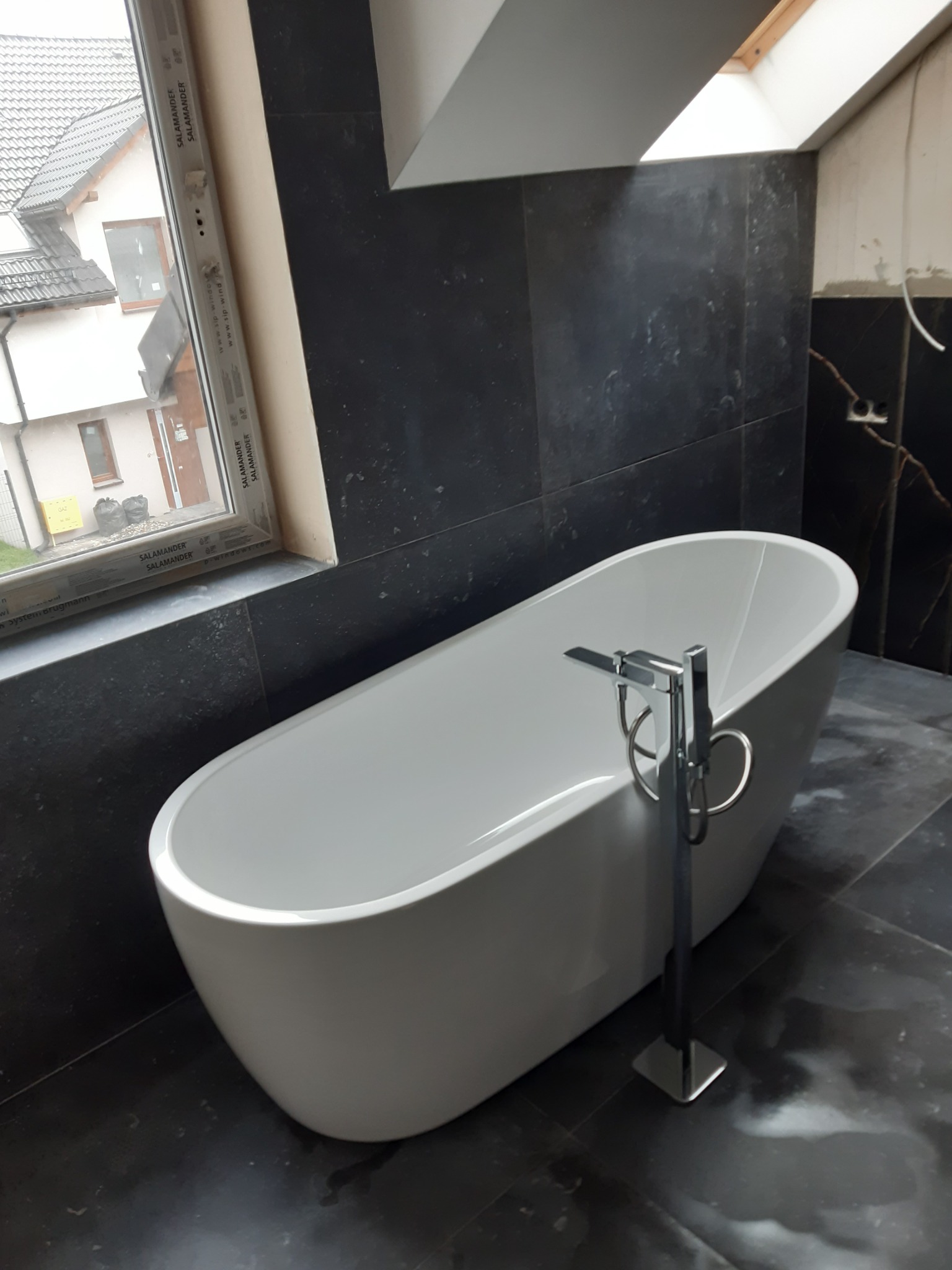 Nowoczesna, wolnostojąca wanna z chromowaną baterią w łazience z czarnymi, marmurkowymi płytkami, widok na okno dachowe i okno z widokiem na dom sąsiada.