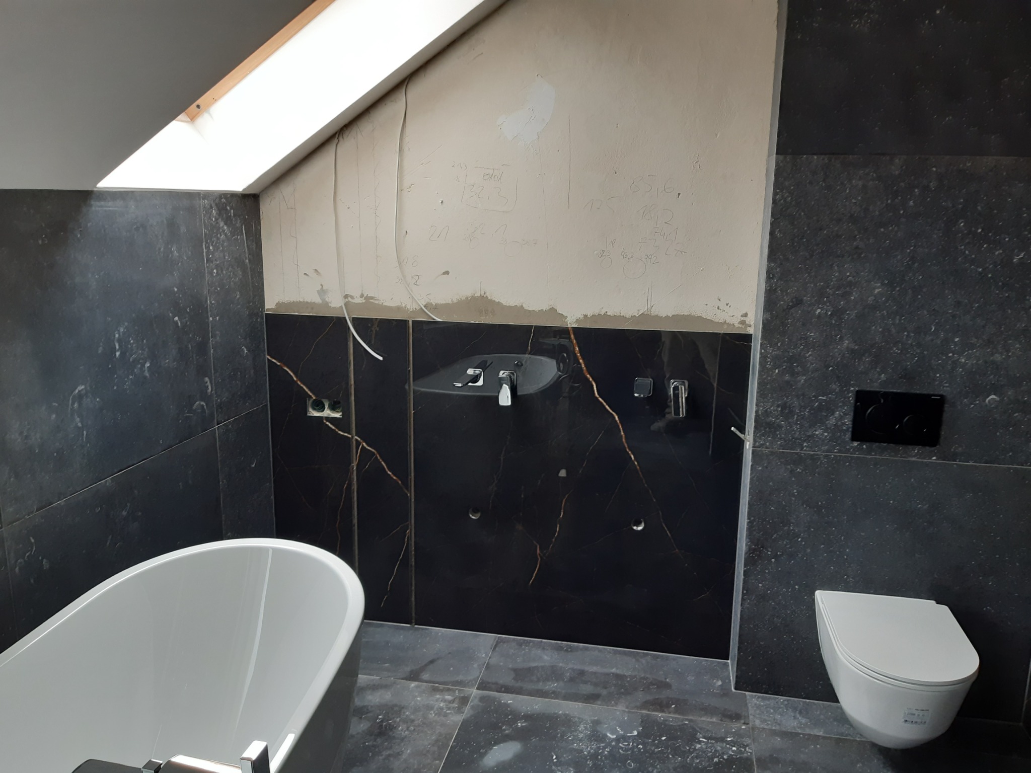 Łazienka w trakcie remontu w poddaszu: czarne płytki z marmurowym wzorem na ścianach i podłodze, biała wanna wolnostojąca, wisząca toaleta, umywalka, ściana z instalacjami i notatkami budowlanymi.