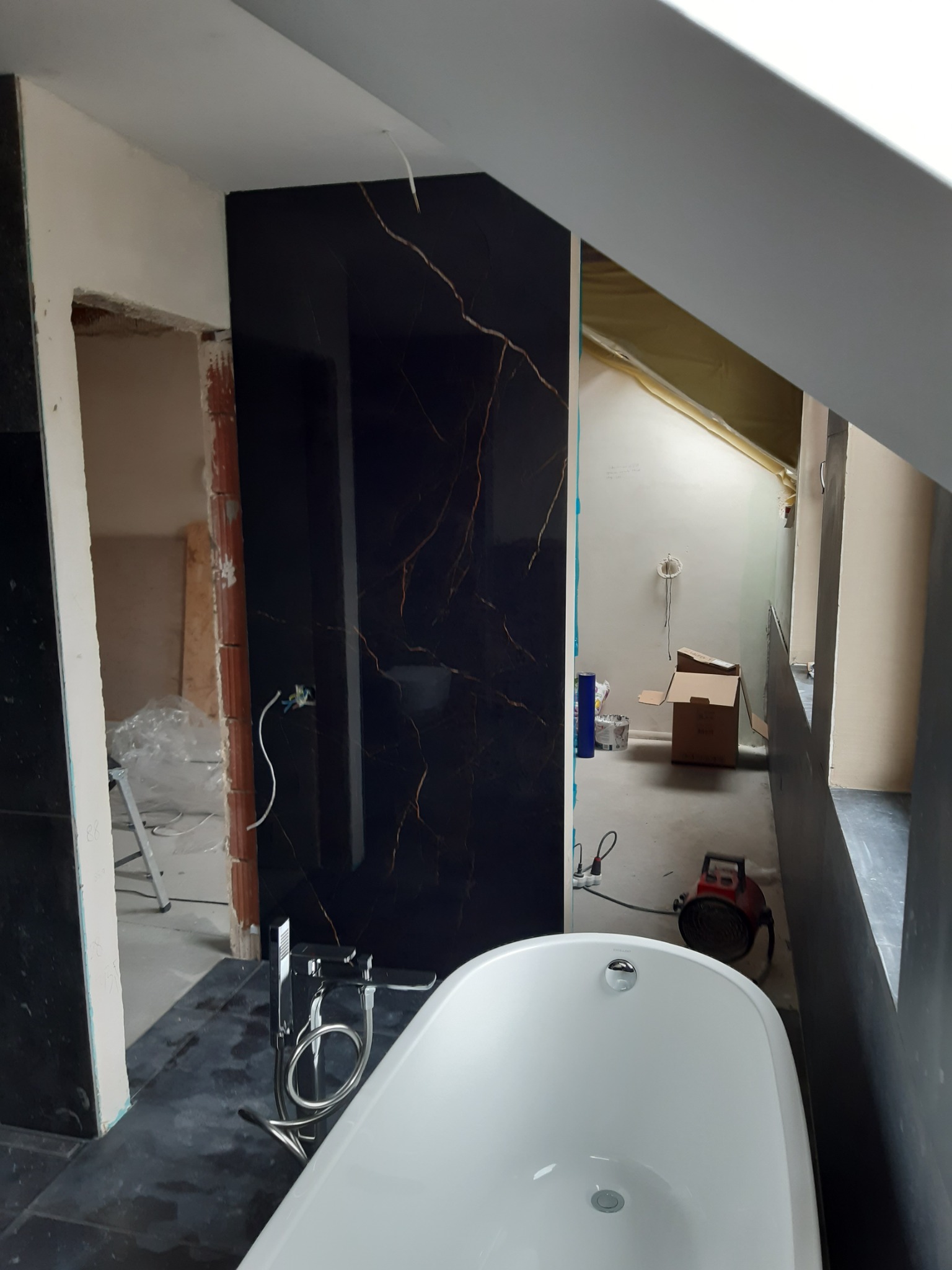 Łazienka w trakcie remontu: czarne płytki imitujące marmur z złotymi żyłami na ścianie, biała wanna wolnostojąca z chromowaną baterią, widoczne niedokończone ściany i narzędzia.
