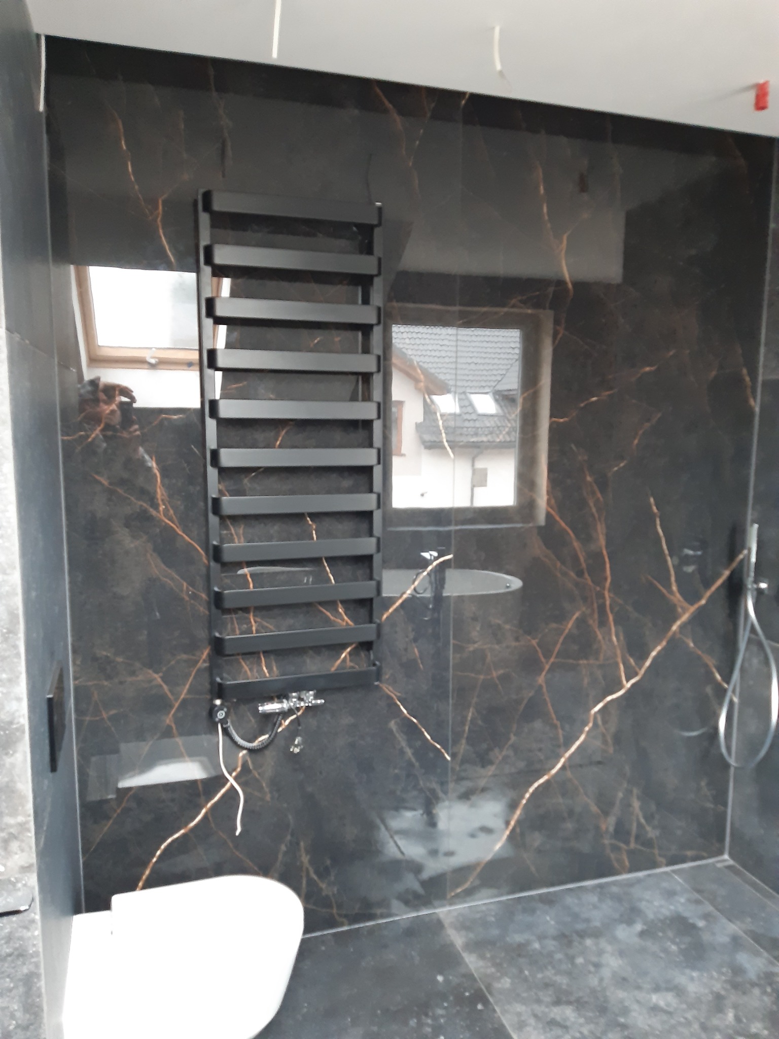 Nowoczesna łazienka z czarnymi płytkami imitującymi marmur z złotymi żyłami, grzejnik drabinkowy w kolorze czarnym, biała toaleta w rogu i widoczne elementy instalacji prysznicowej.