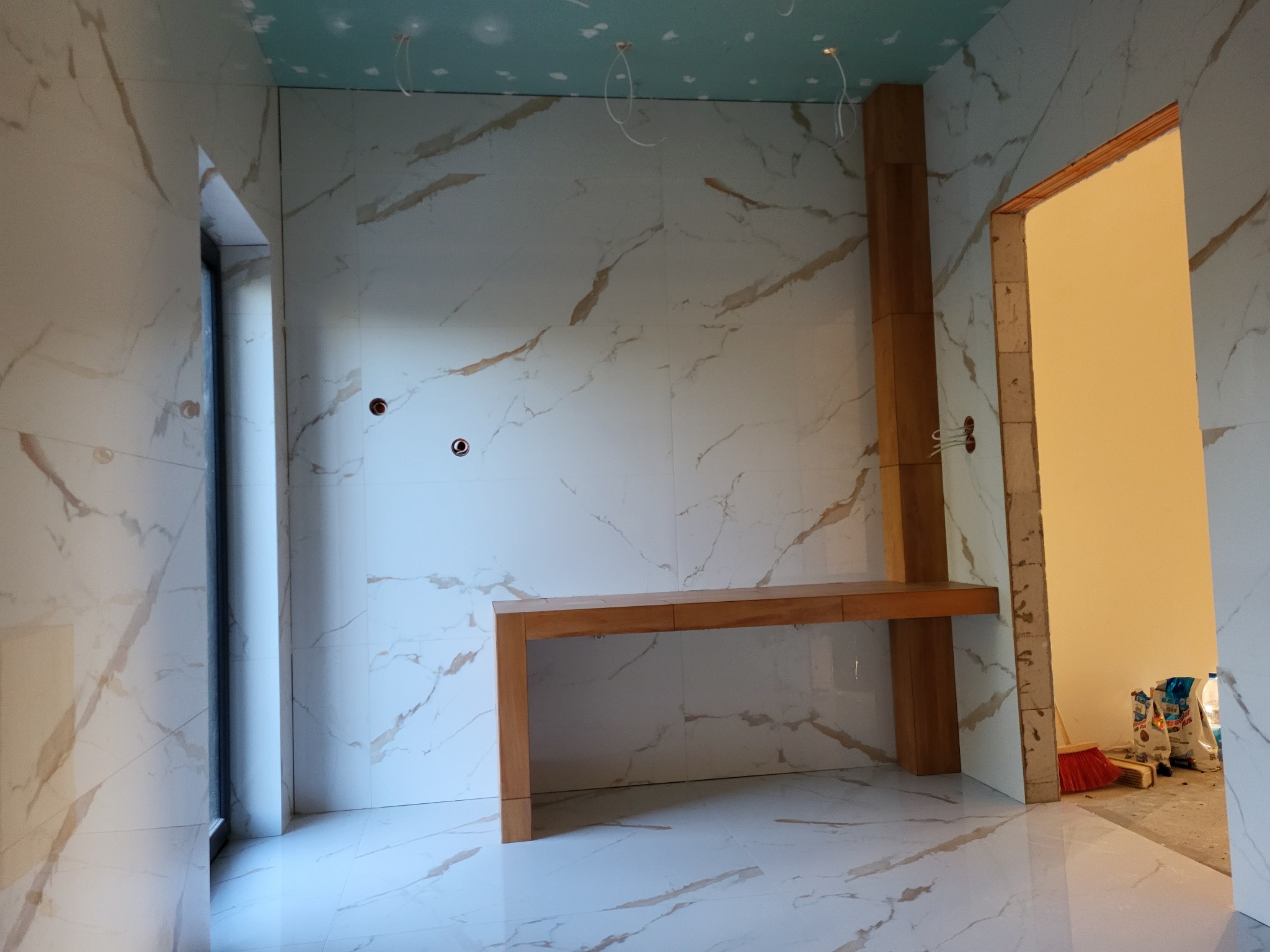 Niedokończona łazienka z marmurowymi płytkami na ścianach i podłodze, drewnianym blatem pod oknem, surowym sufitem z wystającymi kablami i otwartymi drzwiami do sąsiedniego pomieszczenia z workami...