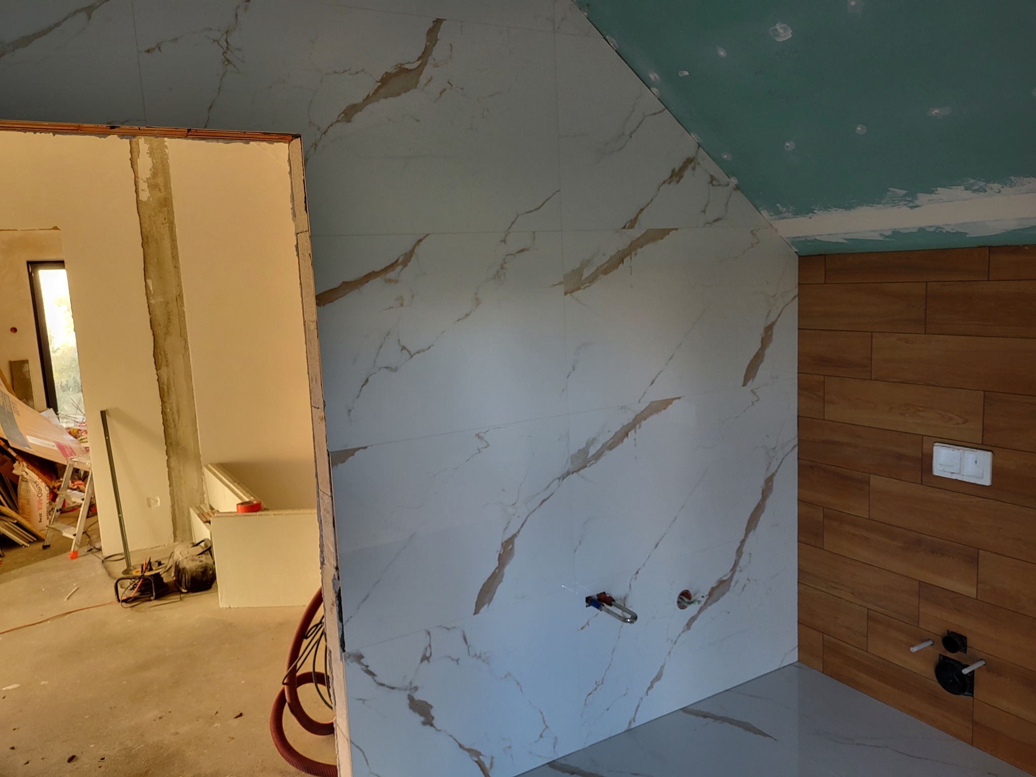 Wnętrze łazienki w trakcie wykańczania: białe płytki imitujące marmur na ścianie, drewnopodobne panele ścienne, niebieski sufit z widocznymi punktami mocowania, oraz widoczne instalacje wodne...