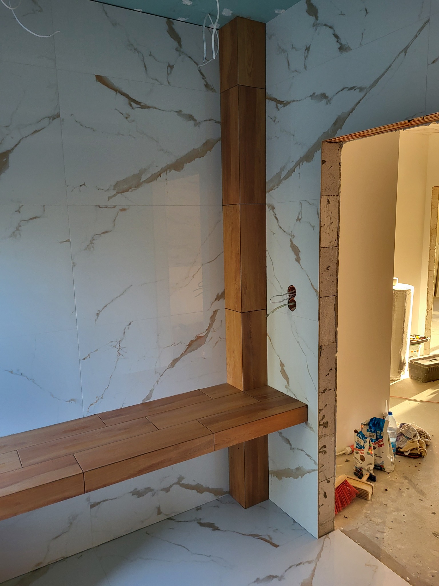 Łazienka w trakcie wykańczania: białe płytki imitujące marmur na ścianach i podłodze, drewniana półka i obudowa pionowa, widoczne kable elektryczne, surowy otwór drzwiowy, worki z zaprawą...