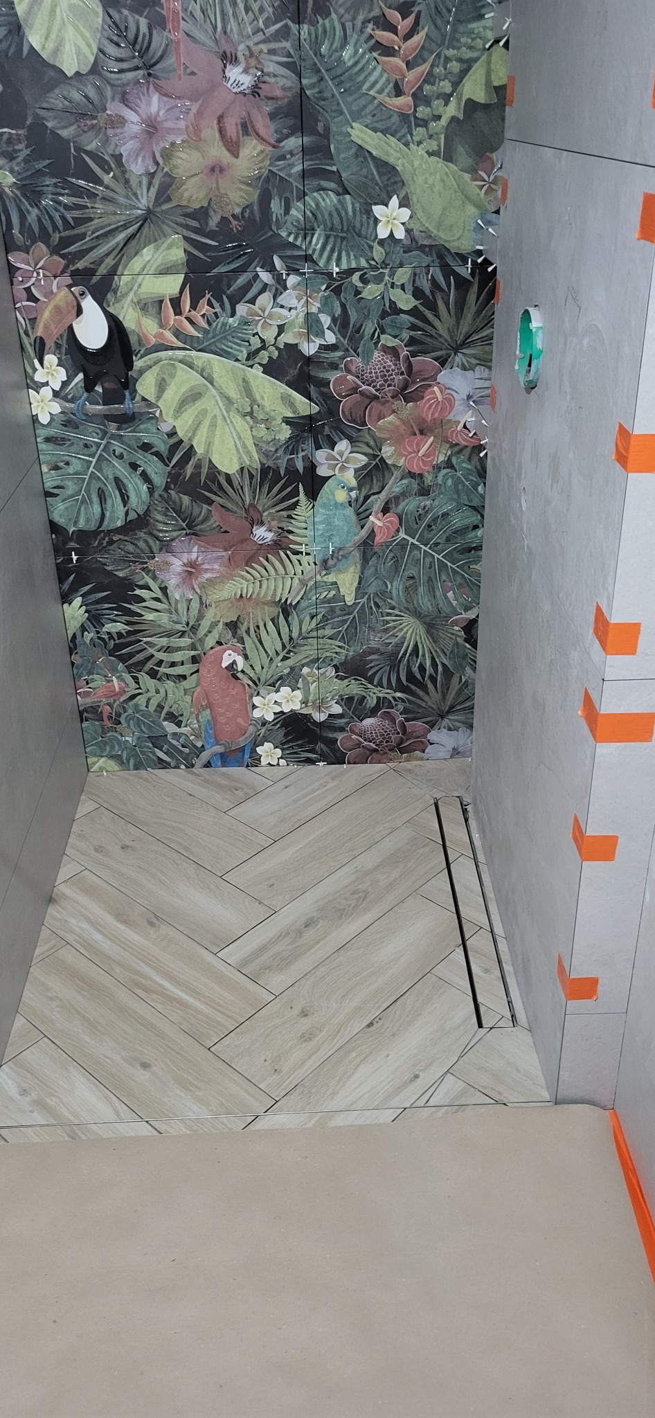 Wykończona kabina prysznicowa z płytkami imitującymi drewno ułożonymi w jodełkę na podłodze i płytkami z motywem tropikalnych roślin i zwierząt na ścianach, widoczne pomarańczowe kliny poziomujące.