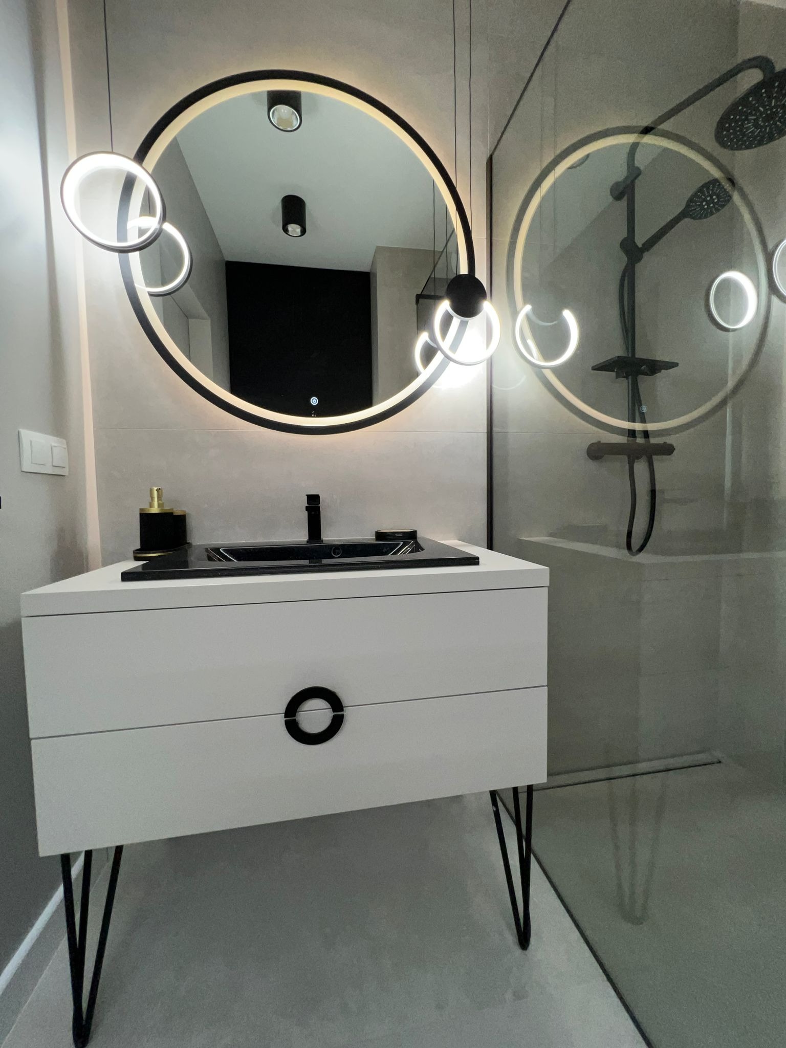 Nowoczesna łazienka z białą szafką pod umywalkę na czarnych nóżkach, czarnym blatem i okrągłym lustrem podświetlanym LED, z kabiną prysznicową w tle.