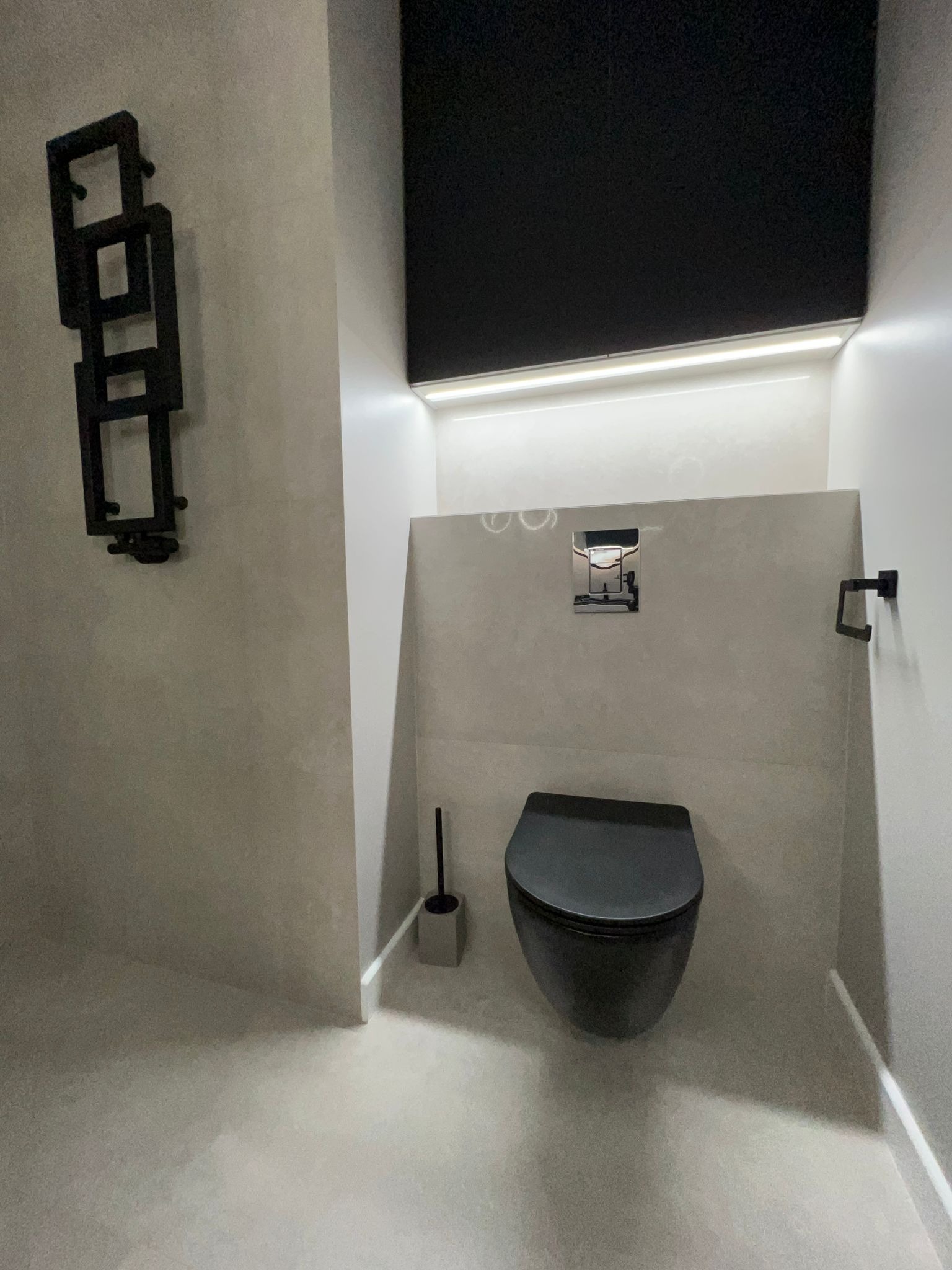 Minimalistyczna łazienka w odcieniach szarości z podwieszaną toaletą w kolorze czarnym, dekoracyjnym grzejnikiem drabinkowym i ukrytym oświetleniem LED.