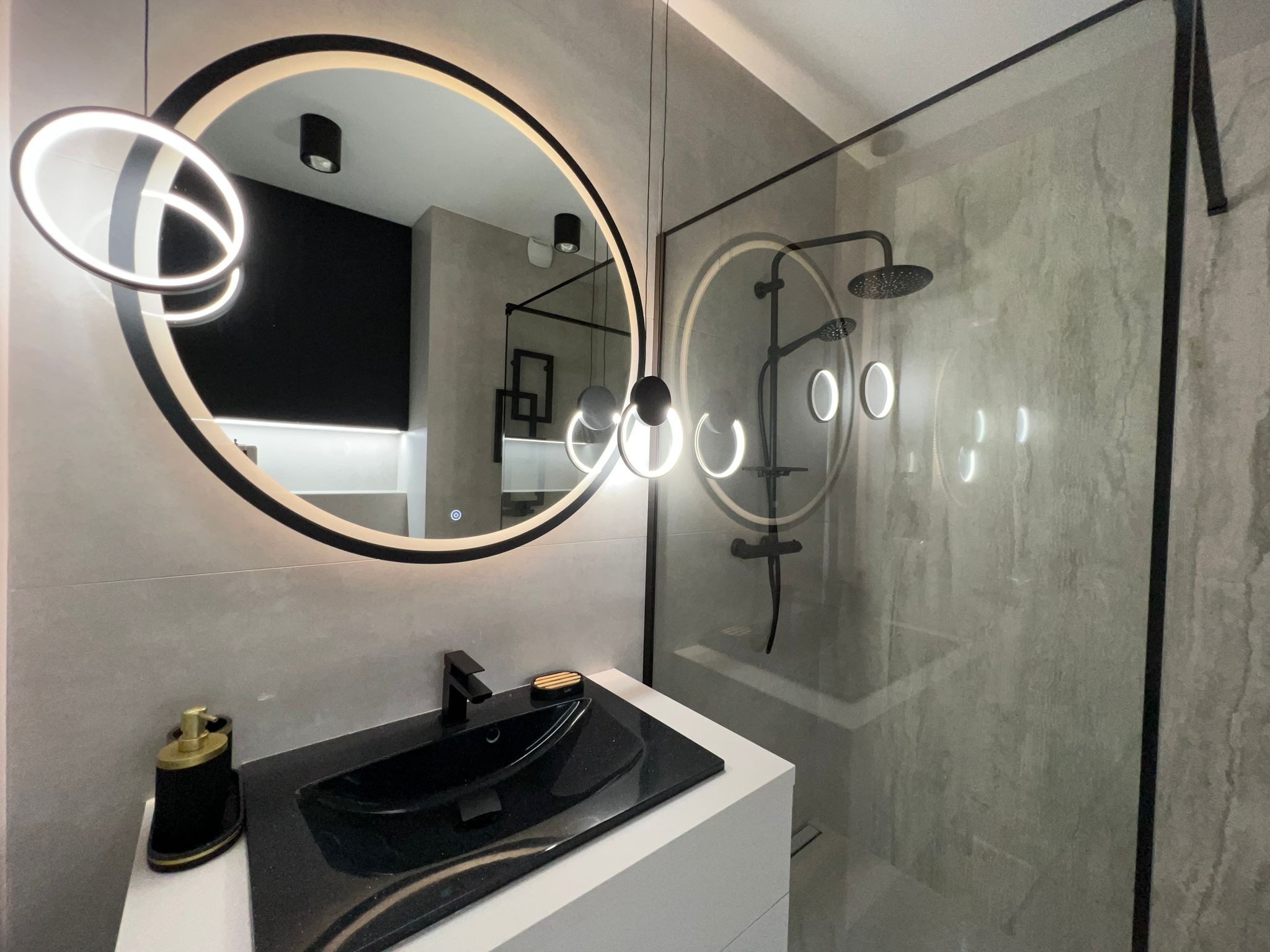Nowoczesna łazienka z czarną armaturą, owalnym lustrem z podświetleniem LED i szklaną kabiną prysznicową z deszczownicą, całość utrzymana w szaro-białej kolorystyce.