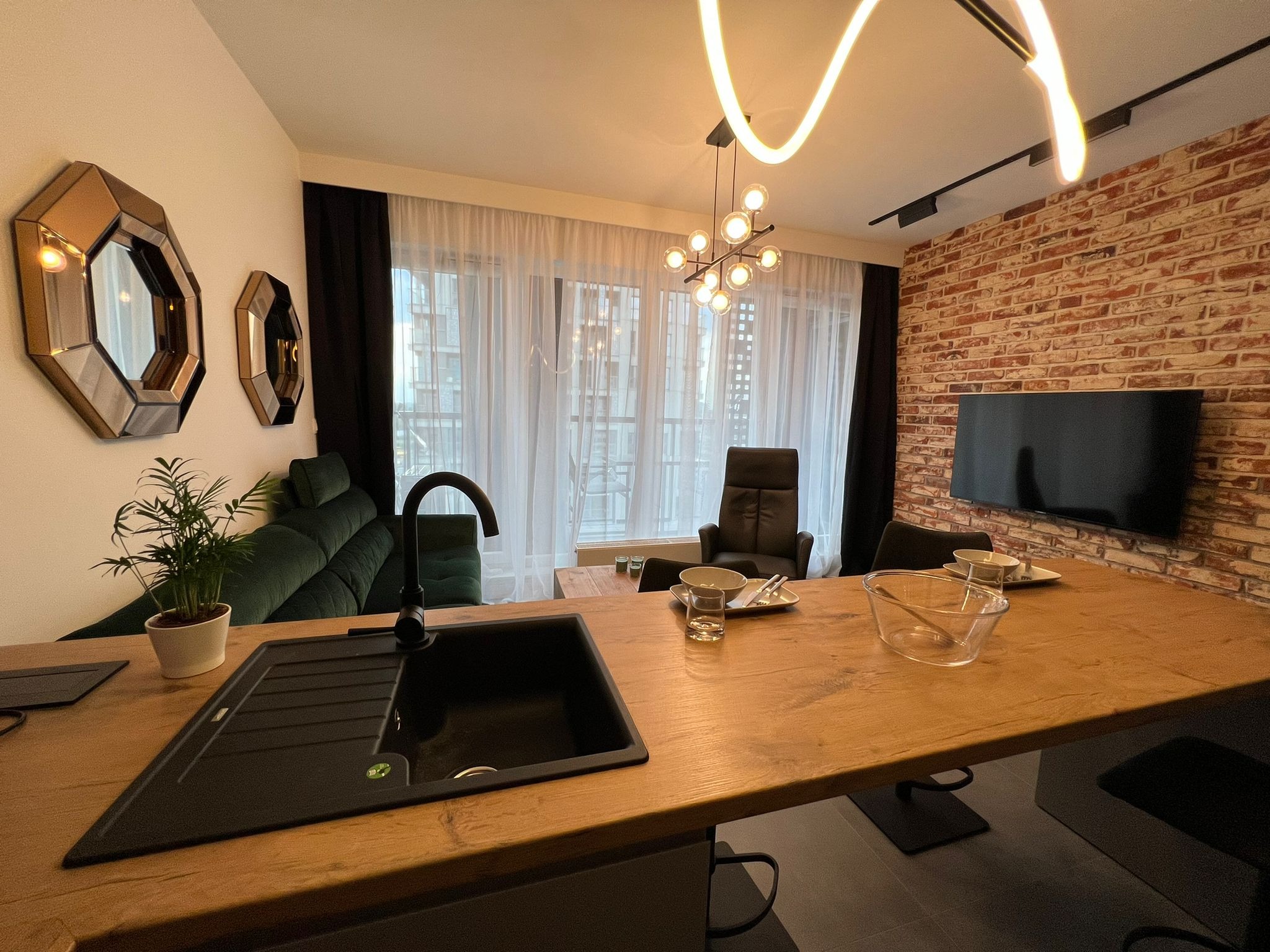 Nowoczesna aranżacja wnętrza z czarnym, narożnym zlewem w drewnianym blacie, zieloną sofą i ścianą z cegły w tle, oświetlona designerskimi lampami.