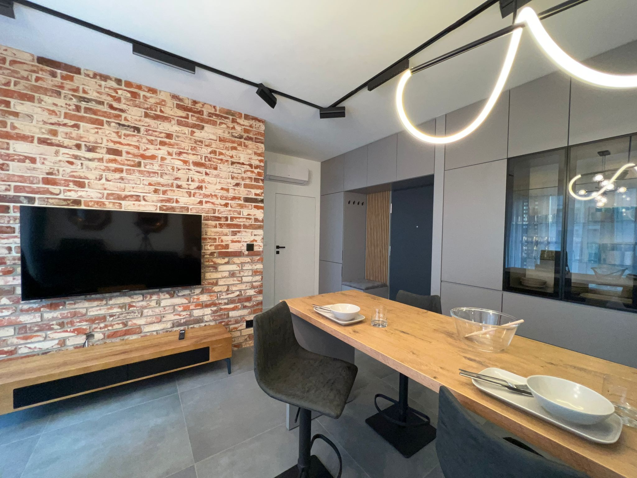 Nowoczesne wnętrze mieszkania z ceglaną ścianą, drewnianym stolikiem kawowym pod telewizorem, szarymi szafkami, oświetleniem punktowym i wiszącą lampą LED w kształcie fali.
