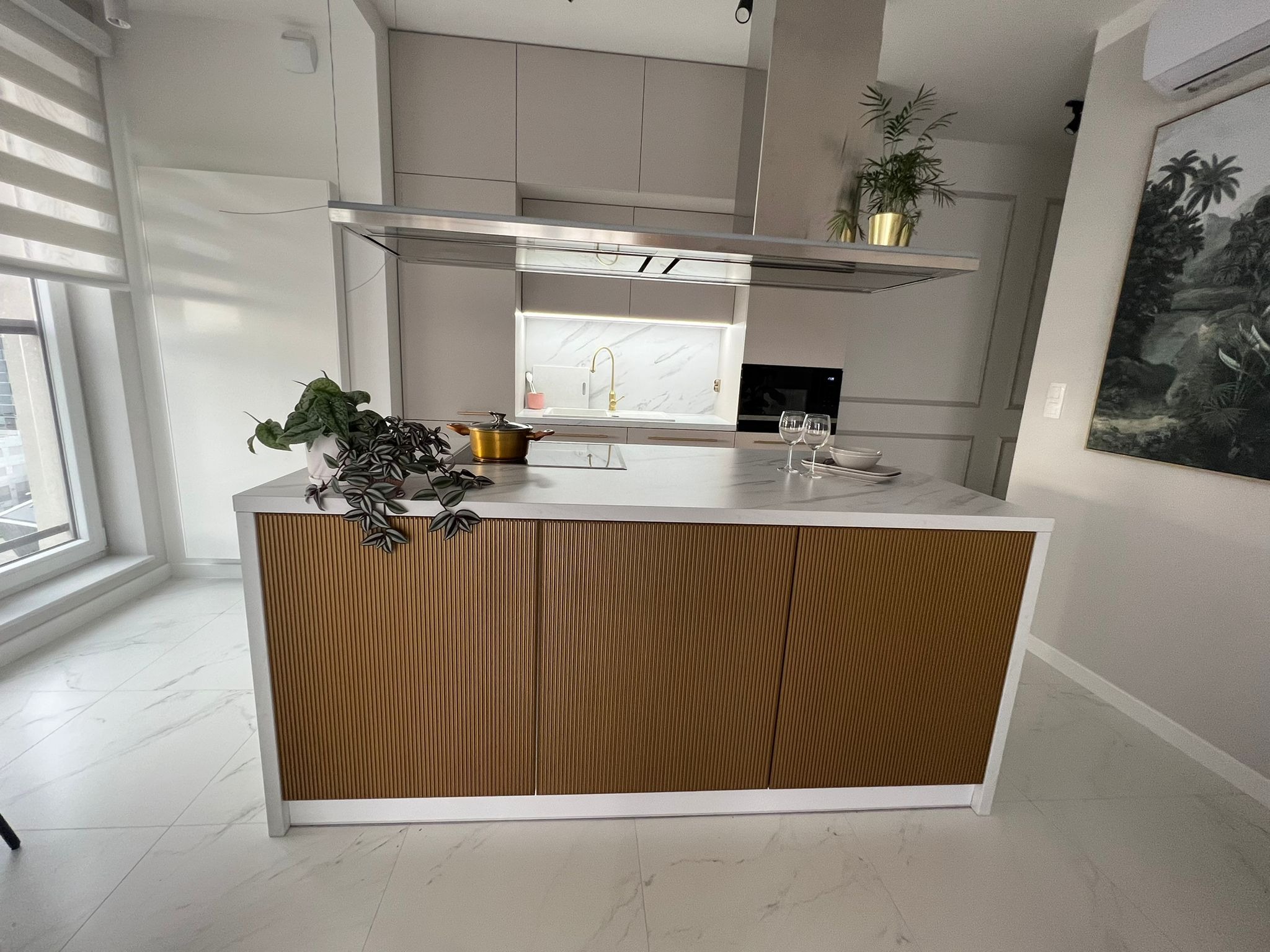 Nowoczesna kuchnia z wyspą o ryflowanych, brązowych frontach, marmurowym blatem, złotą armaturą i roślinami doniczkowymi jako elementami dekoracyjnymi.