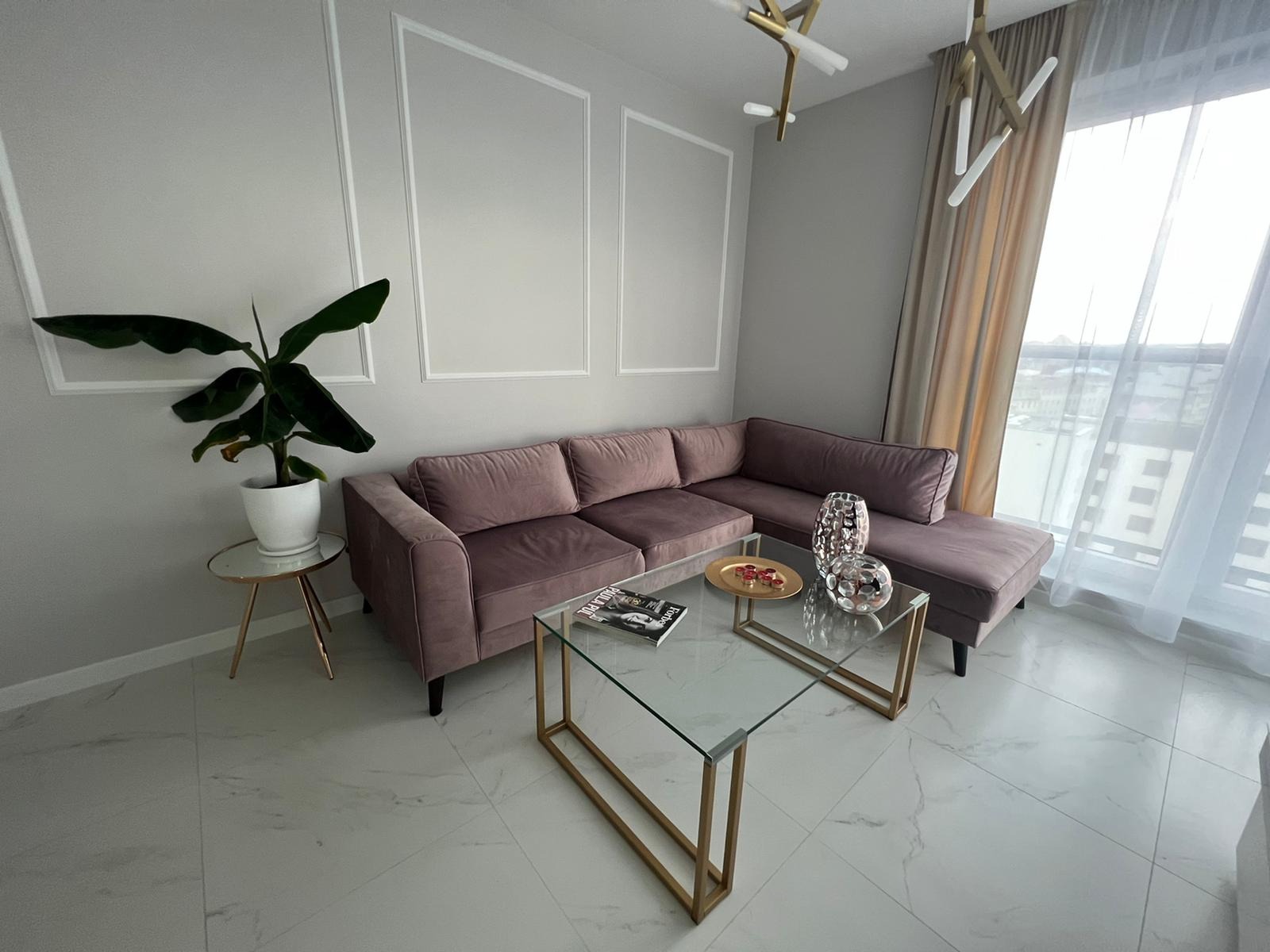 Elegancki salon z narożną sofą w odcieniach różu, szklanym stolikiem kawowym na złotych nogach, marmurową podłogą i dekoracyjnymi listwami ściennymi.