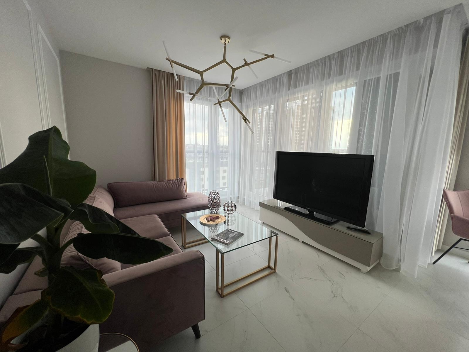 Jasne wnętrze salonu z marmurową podłogą, narożną sofą w odcieniach różu, szklanym stolikiem kawowym z dekoracjami, telewizorem na szafce RTV i nowoczesnym żyrandolem.