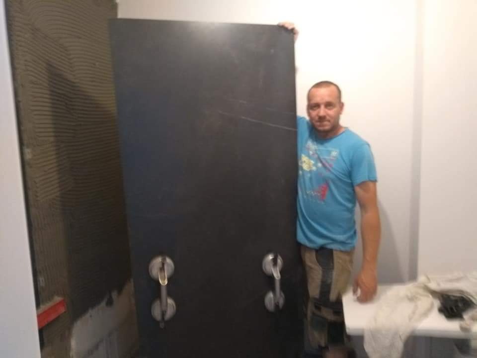 Mężczyzna w niebieskiej koszulce prezentuje dużą, ciemną płytę z przyssawkami, prawdopodobnie płytę gresową, na tle przygotowanej do położenia płytek ściany w łazience.